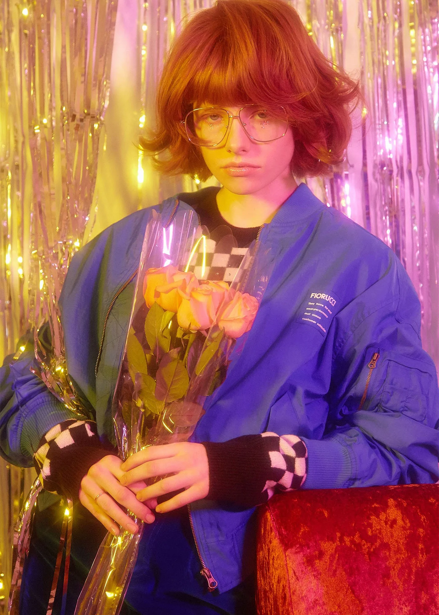 A young person with red hair and glasses holding a bouquet of pink and yellow roses in a decorative setting with pink and gold tinsel background.