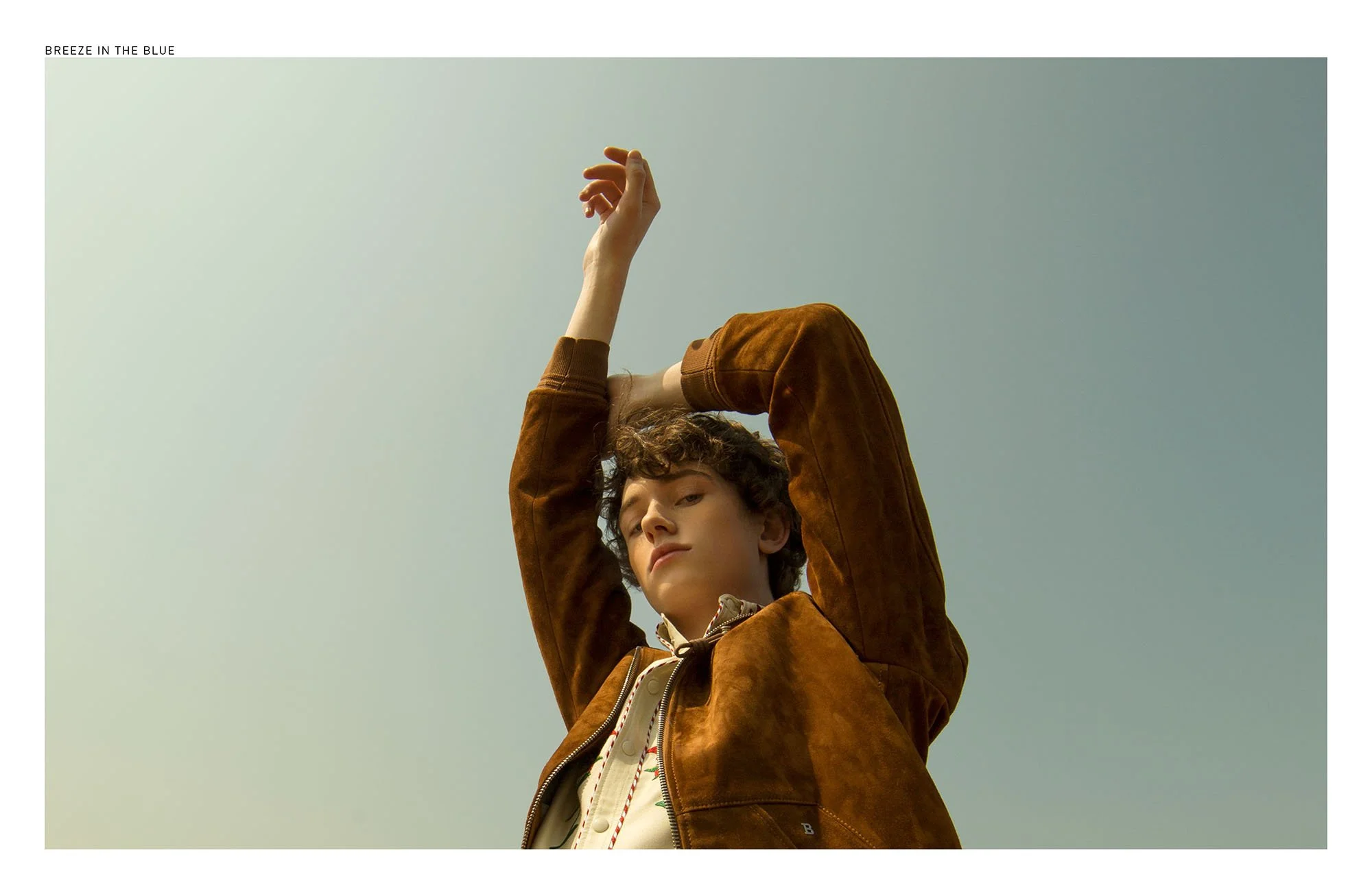 Young person with curly hair, wearing a brown jacket, standing outdoors with arms raised above head against a pale sky.