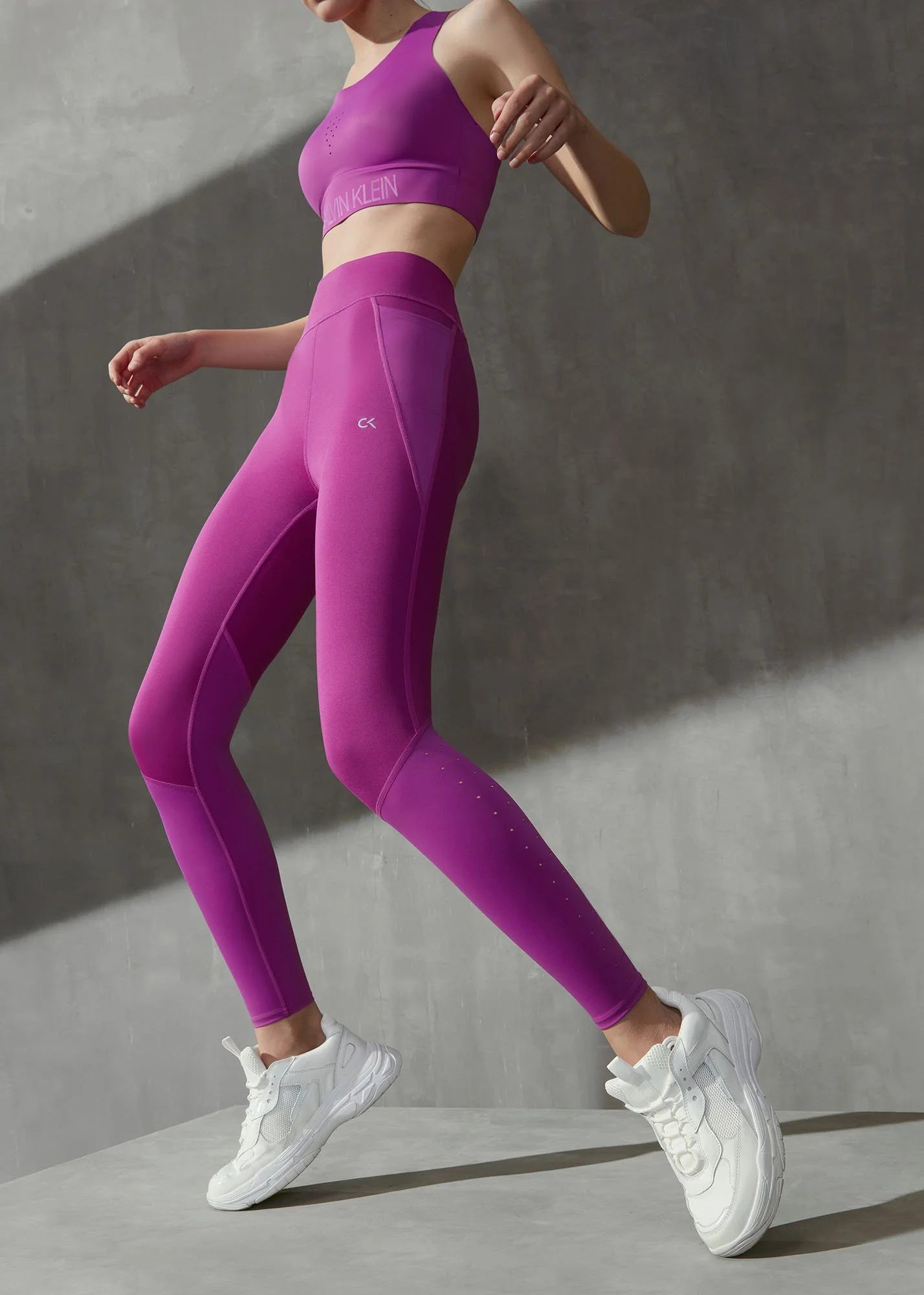 A person wearing a purple athletic crop top and matching leggings, white sneakers, and standing on a light gray floor against a concrete wall.