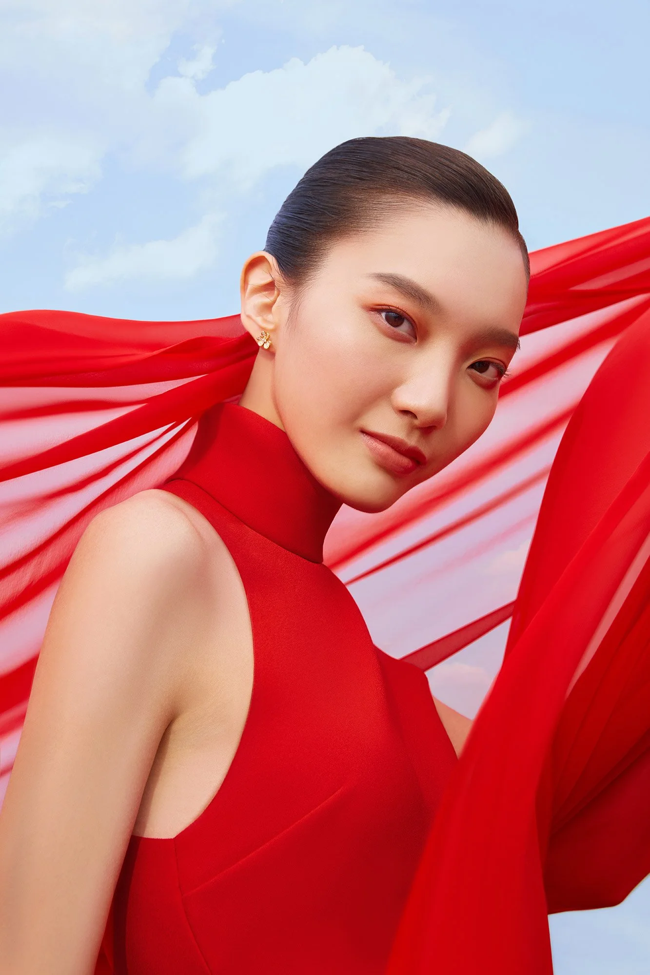 A woman with dark hair in a sleek bun wearing a red sleeveless dress, with matching red fabric flowing in the background, against a partly cloudy sky.