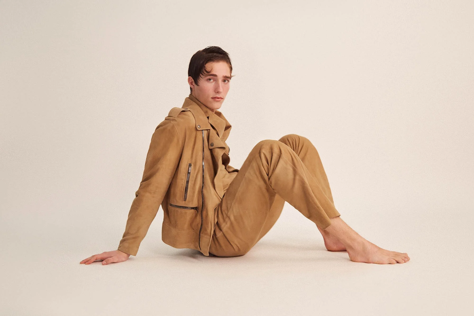A young man sitting on the floor with his back against a plain cream background. He is wearing a tan suede jacket and matching pants, with no shoes.