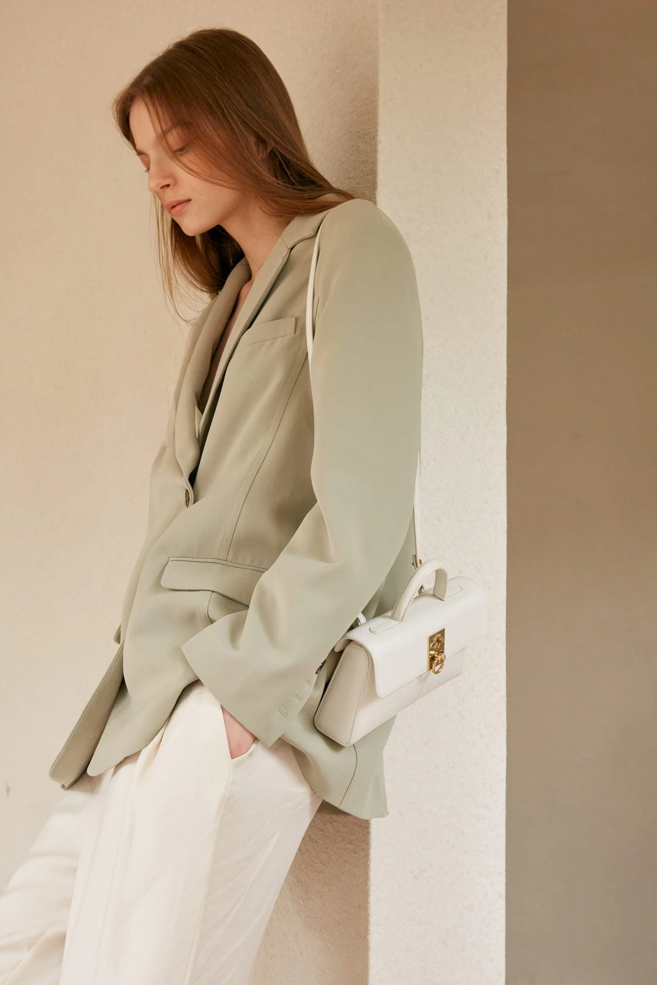 A woman with long brown hair, wearing a light beige blazer and white pants, standing against a beige wall. She has a white handbag with gold hardware hanging from her left arm.