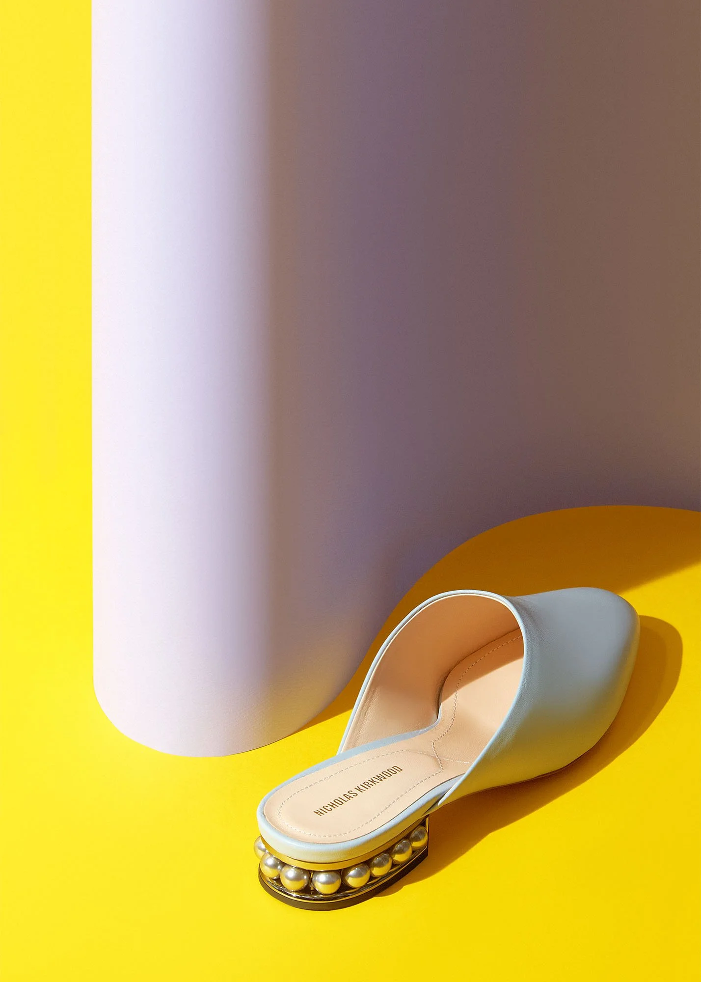 White high-heeled shoe with pearl accents on the heel, placed on a yellow surface with a pink and purple background.