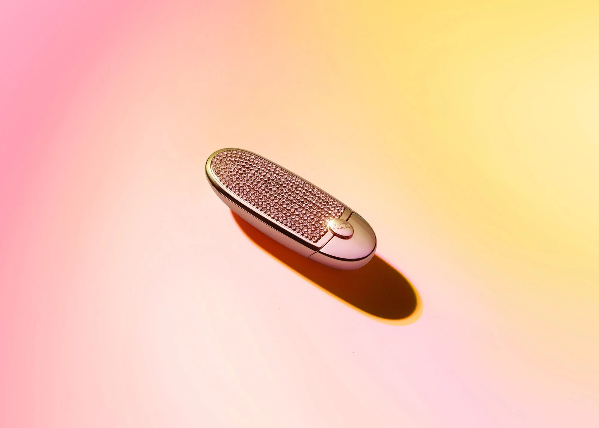 A luxury electric foot massager with a metallic pink and gold finish, decorated with small crystals, placed on a yellow and pink gradient background.