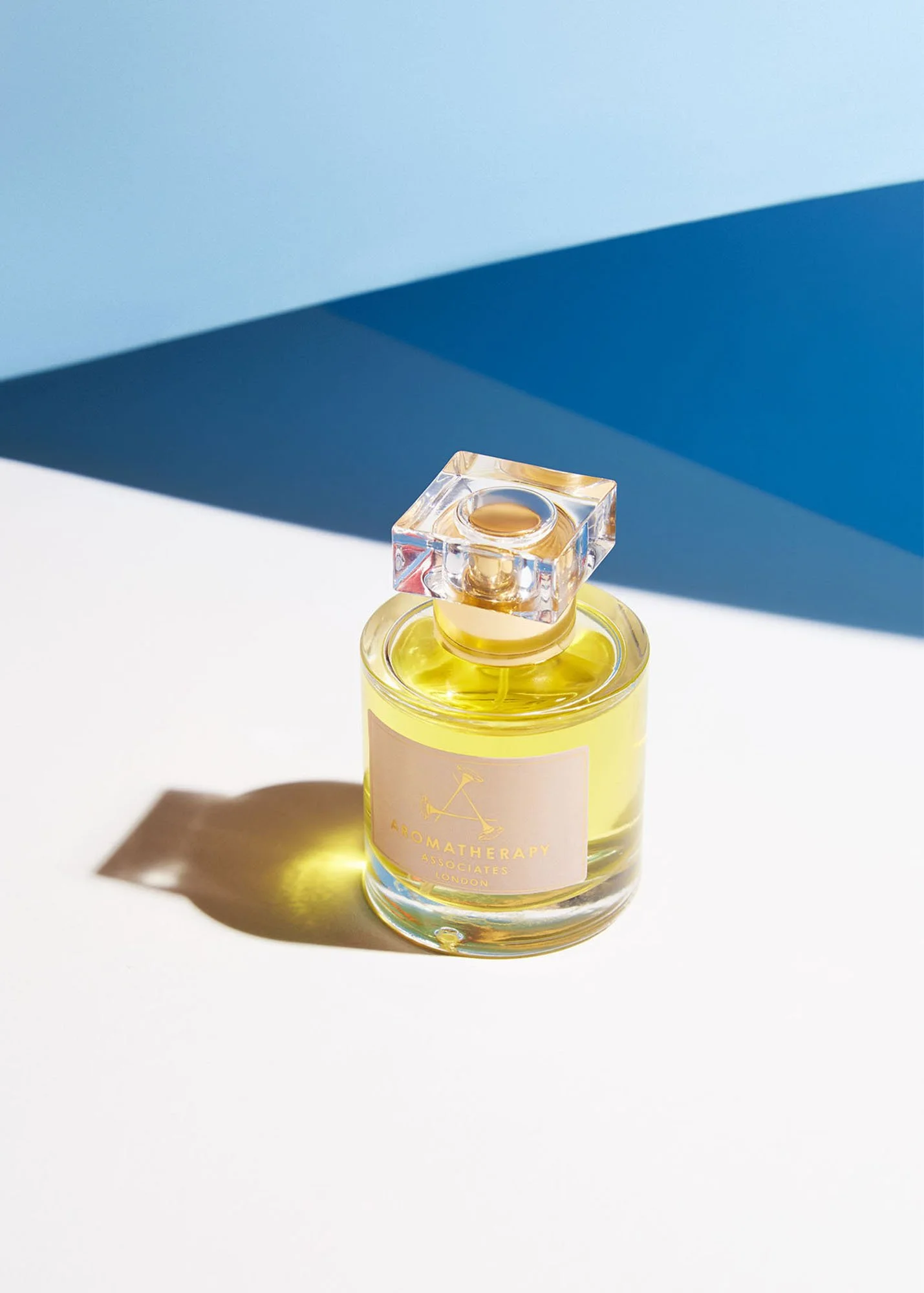 A glass perfume bottle with a yellow liquid and a clear square-shaped cap, sitting on a white surface with a blue background.