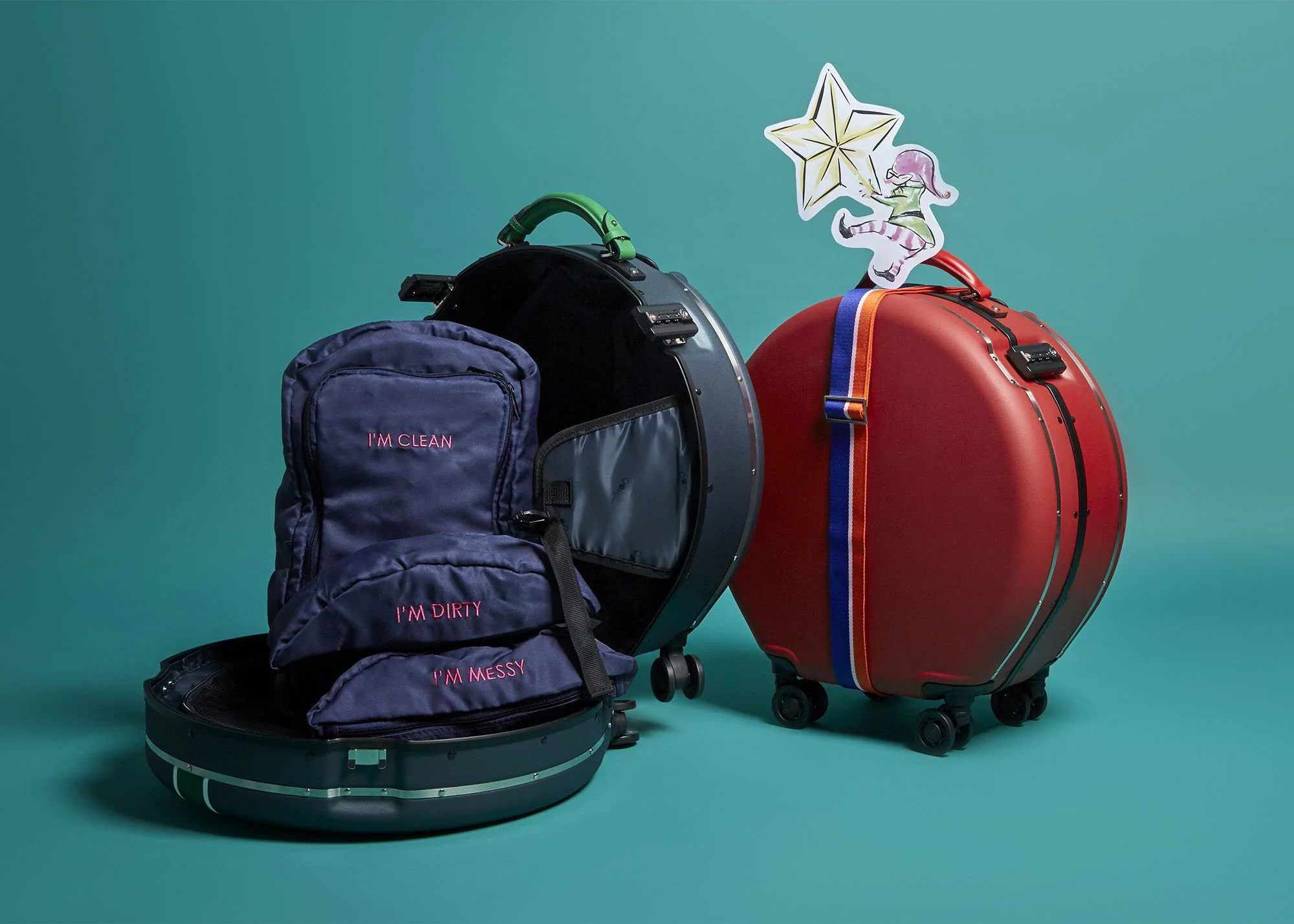 Open suitcase with navy backpack inside, next to a red hard-shell suitcase with a colorful strap, and a paper cutout figure of a girl sitting on a star. The backpacks have embroidered words: 'I'M CLEAN,' 'I'M DIRTY,' and 'I'M MESSY.'