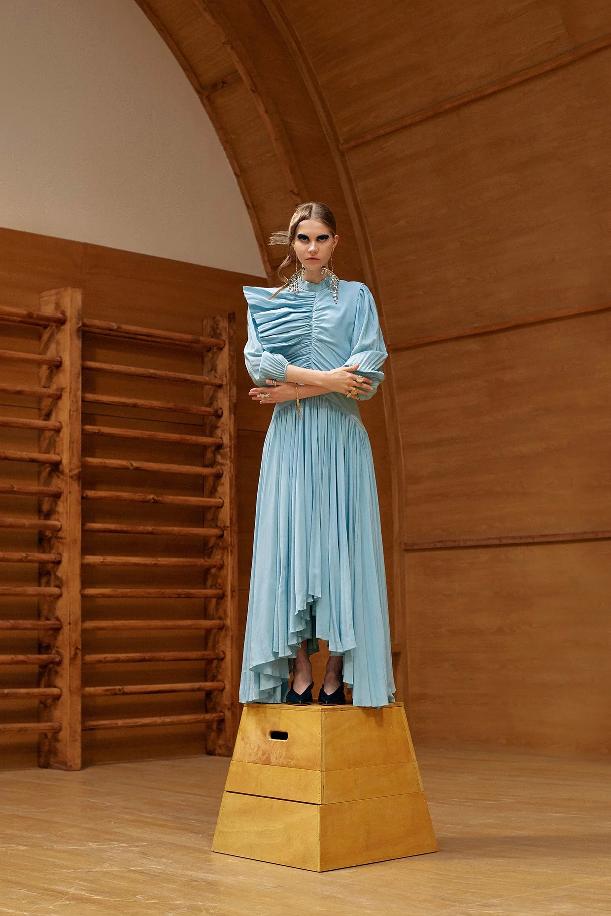 A woman standing on a wooden platform in a wood-paneled room, wearing a long light blue dress with pleats and puffed sleeves, with her arms crossed and looking directly at the camera.