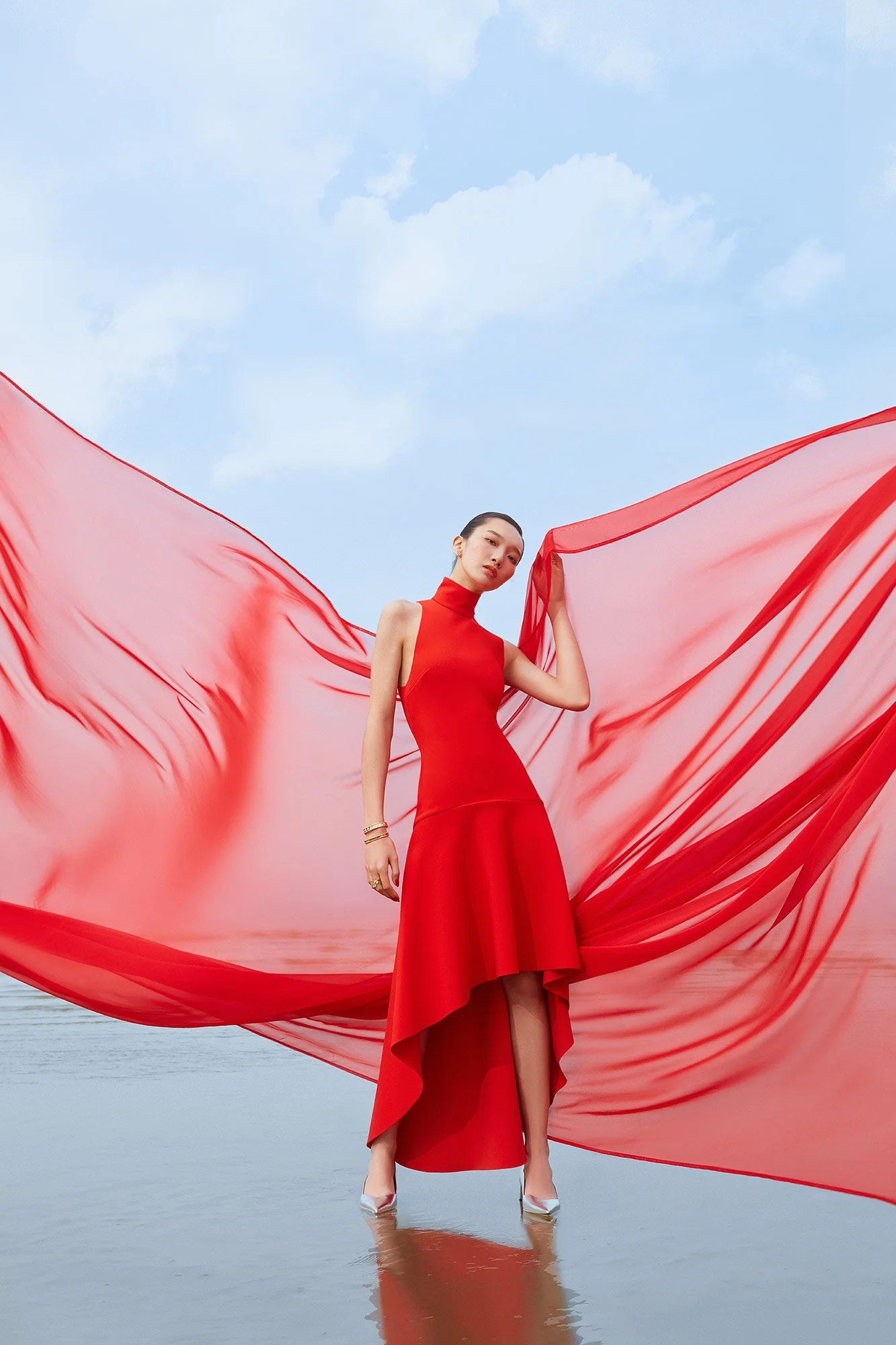 A woman in a red dress standing in water, holding a flowing red fabric against a bright blue sky.