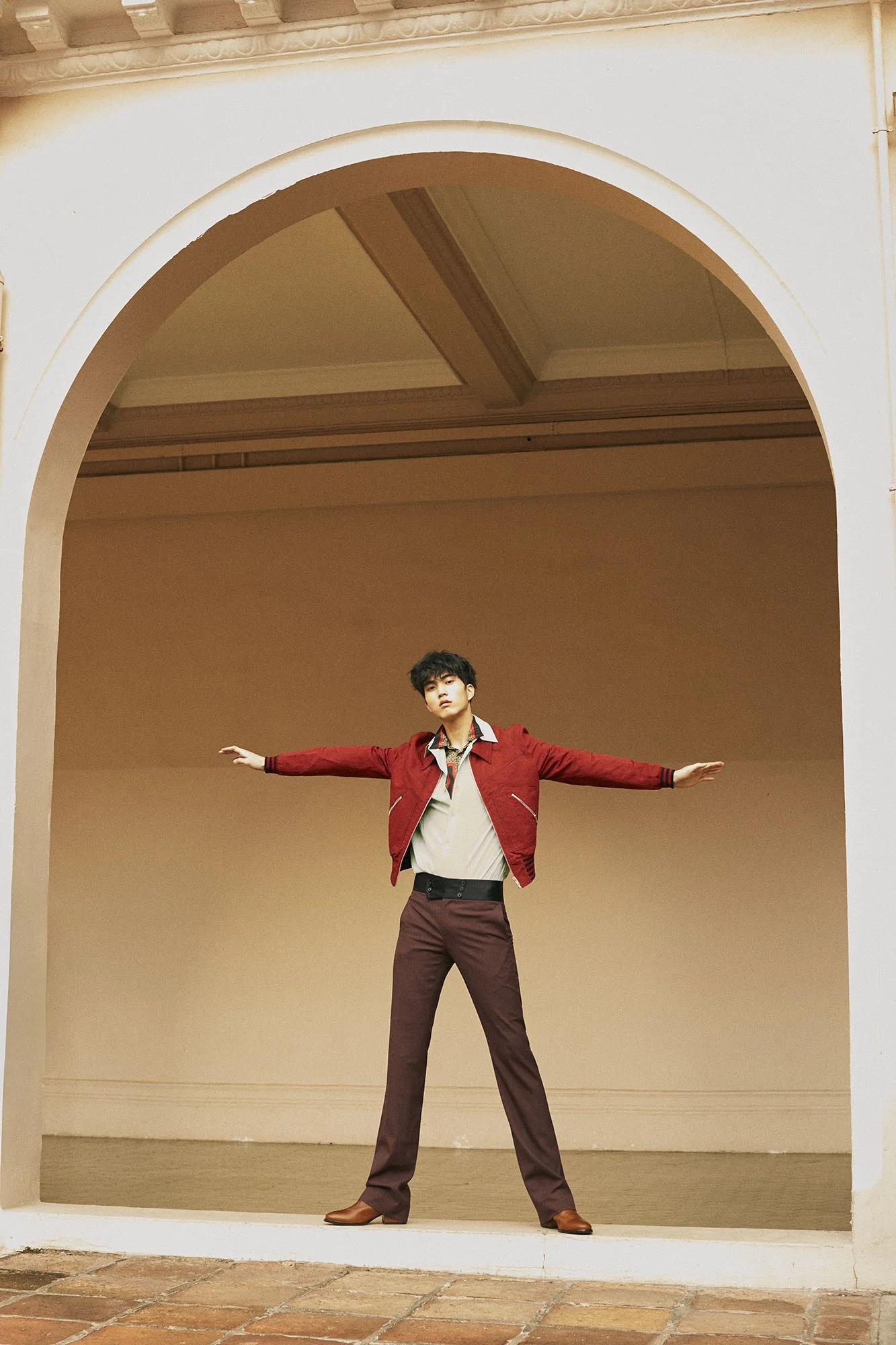 Young man standing with arms outstretched in an archway, dressed in a red jacket, beige shirt, and brown pants.