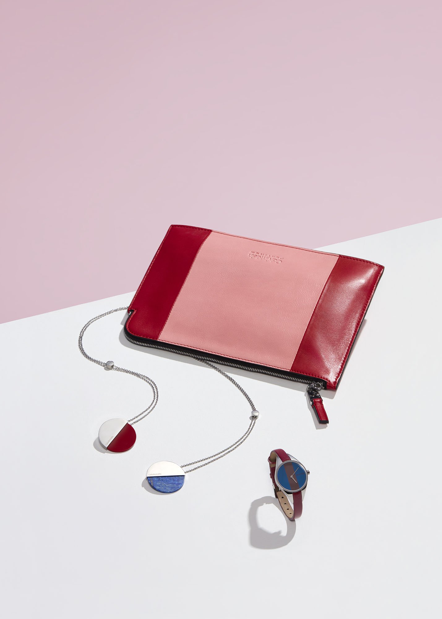 A pink and red leather zippered clutch purse, two colorful round necklaces, and a wristwatch with a red strap are arranged on a surface with a pink and white background.