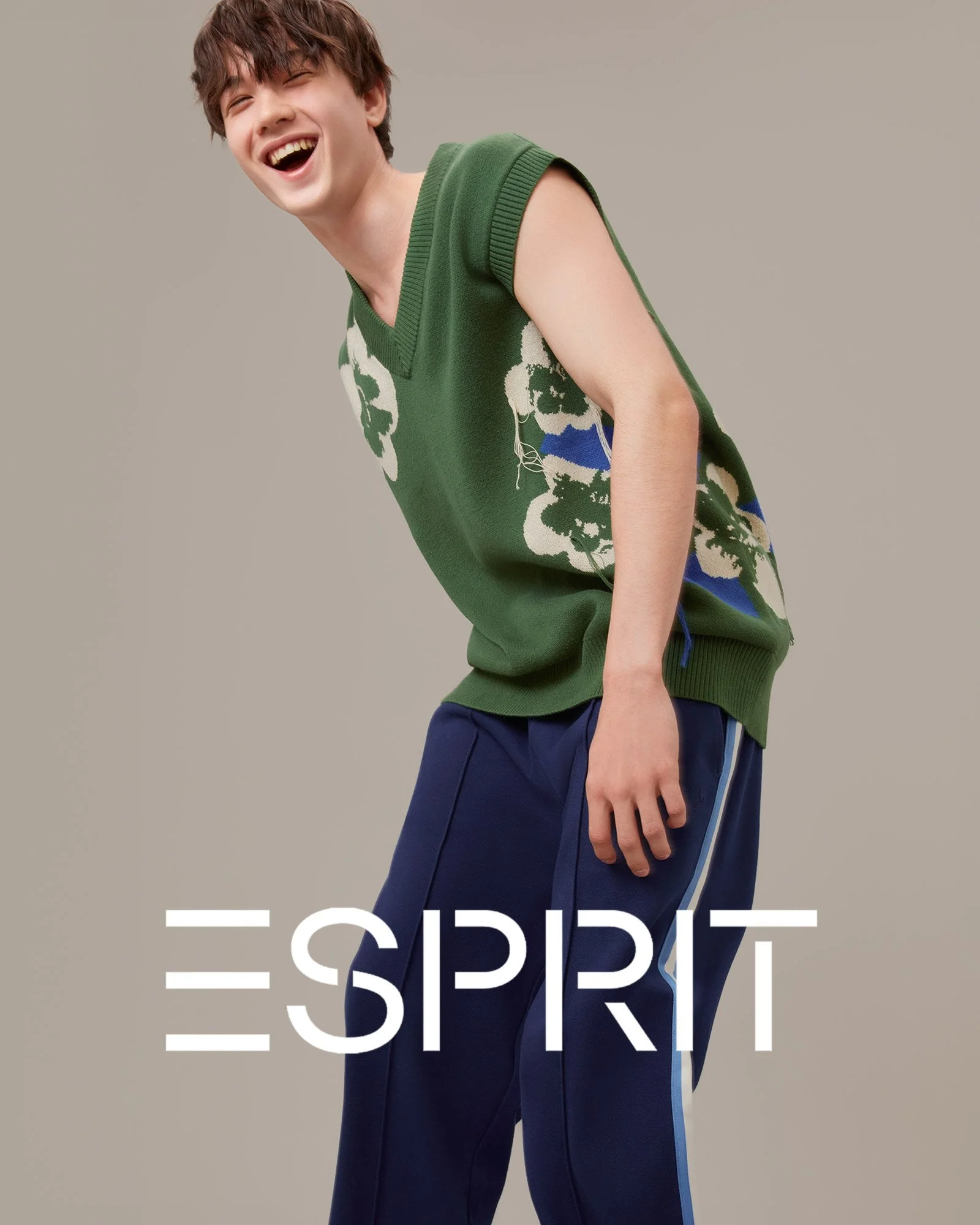 Young man laughing, wearing a green sleeveless sweater vest with a floral pattern and navy blue athletic pants, standing against a neutral background with the Esprit logo at the bottom.
