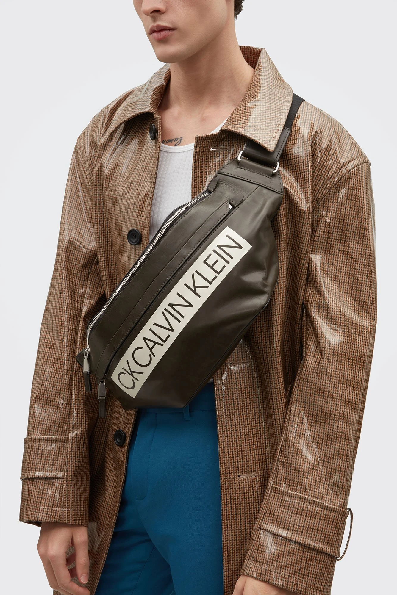A person wearing a brown checkered coat with a white shirt underneath and blue pants, with a black Calvin Klein crossbody bag across their chest.