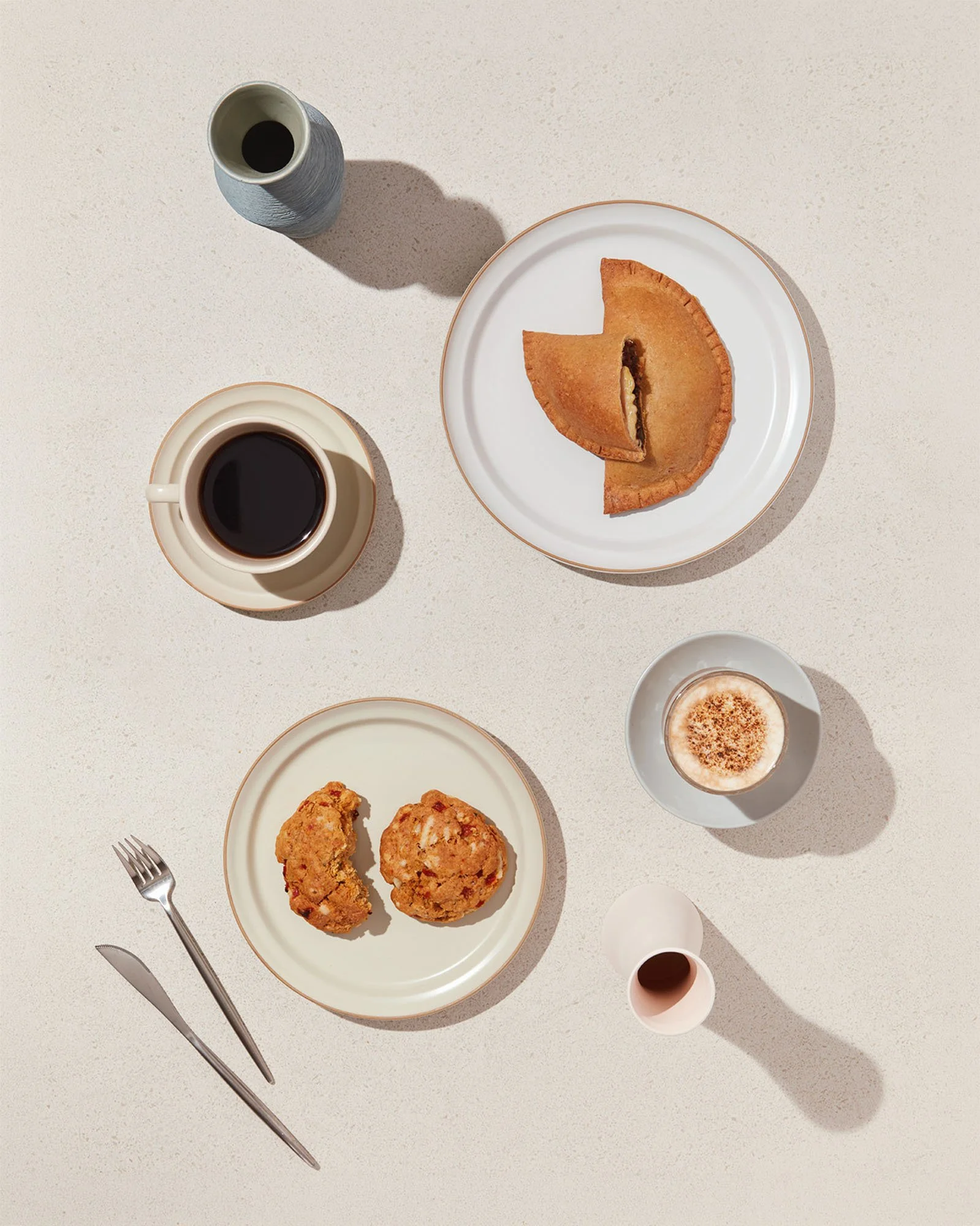 Overhead shot of a breakfast spread including a slice of pie on a white plate, two fried chicken drumsticks on a separate plate, a cup of black coffee, a glass of whiskey, a small cup of whipped dessert with cocoa powder, and a knife and fork on a wh