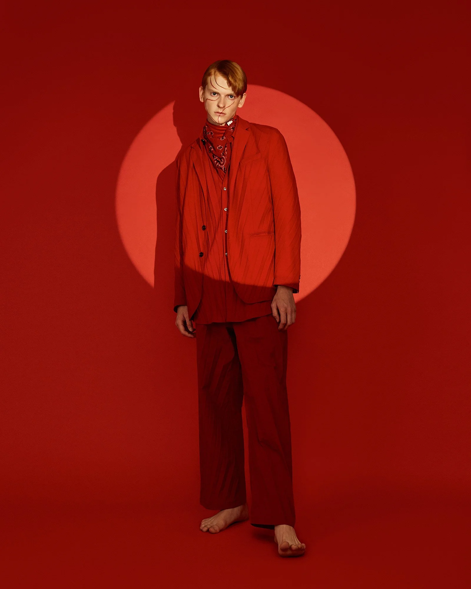 Fashion model wearing a red suit and bandana standing against red background with a circular light or spotlight.