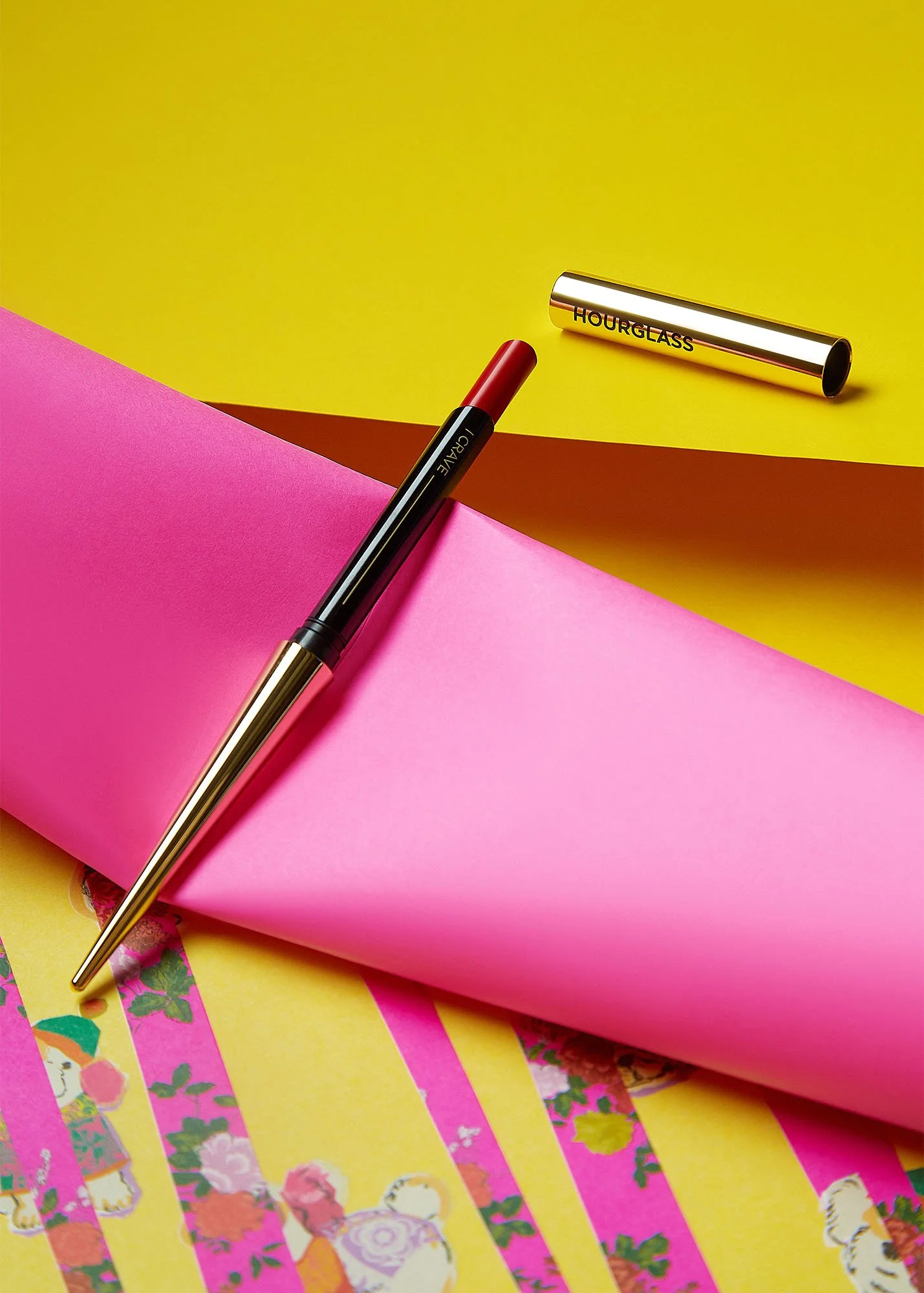 A black and gold pen resting on pink paper, with a gold tube labeled 'HOUR GLASS' nearby on yellow background.