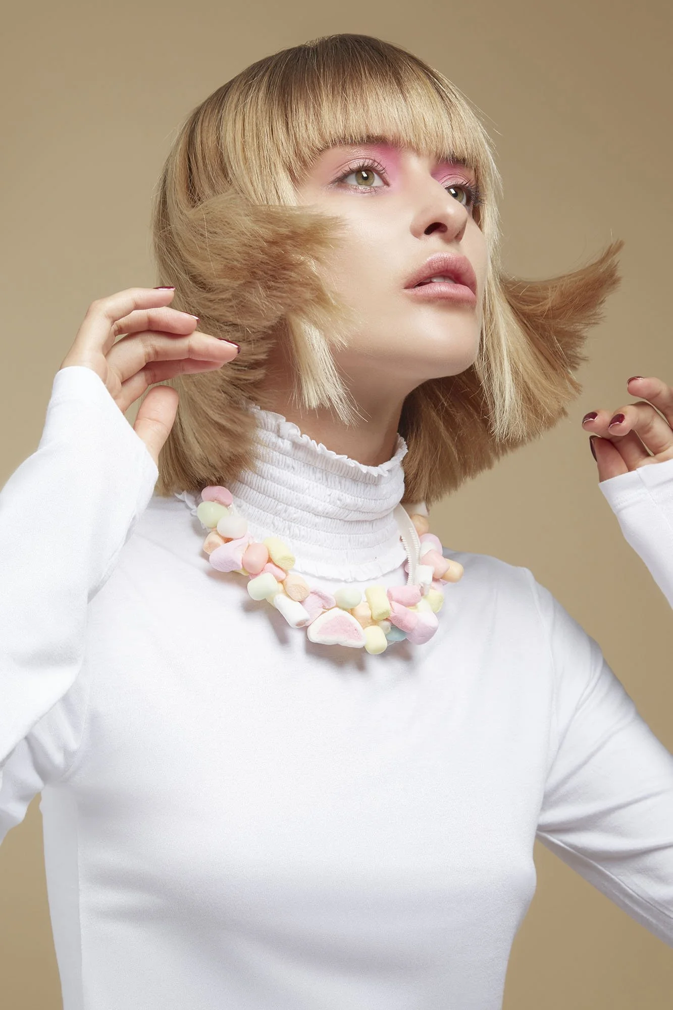 A woman with blonde hair styled in a bob with bangs, wearing a white turtleneck and a colorful marshmallow necklace, poses against a neutral background.