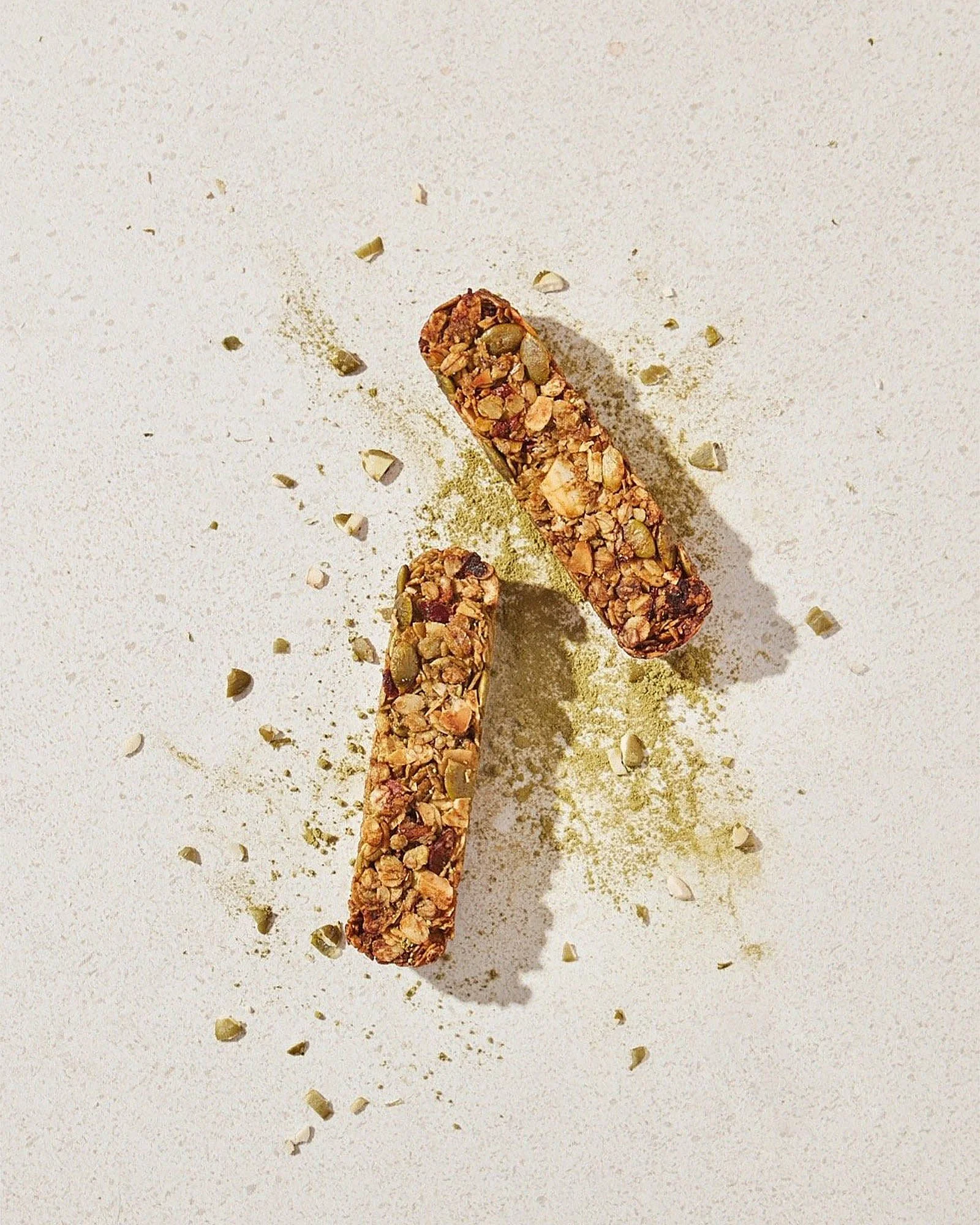Two granola bars with nuts and seeds on a white surface with scattered nuts and green powder.