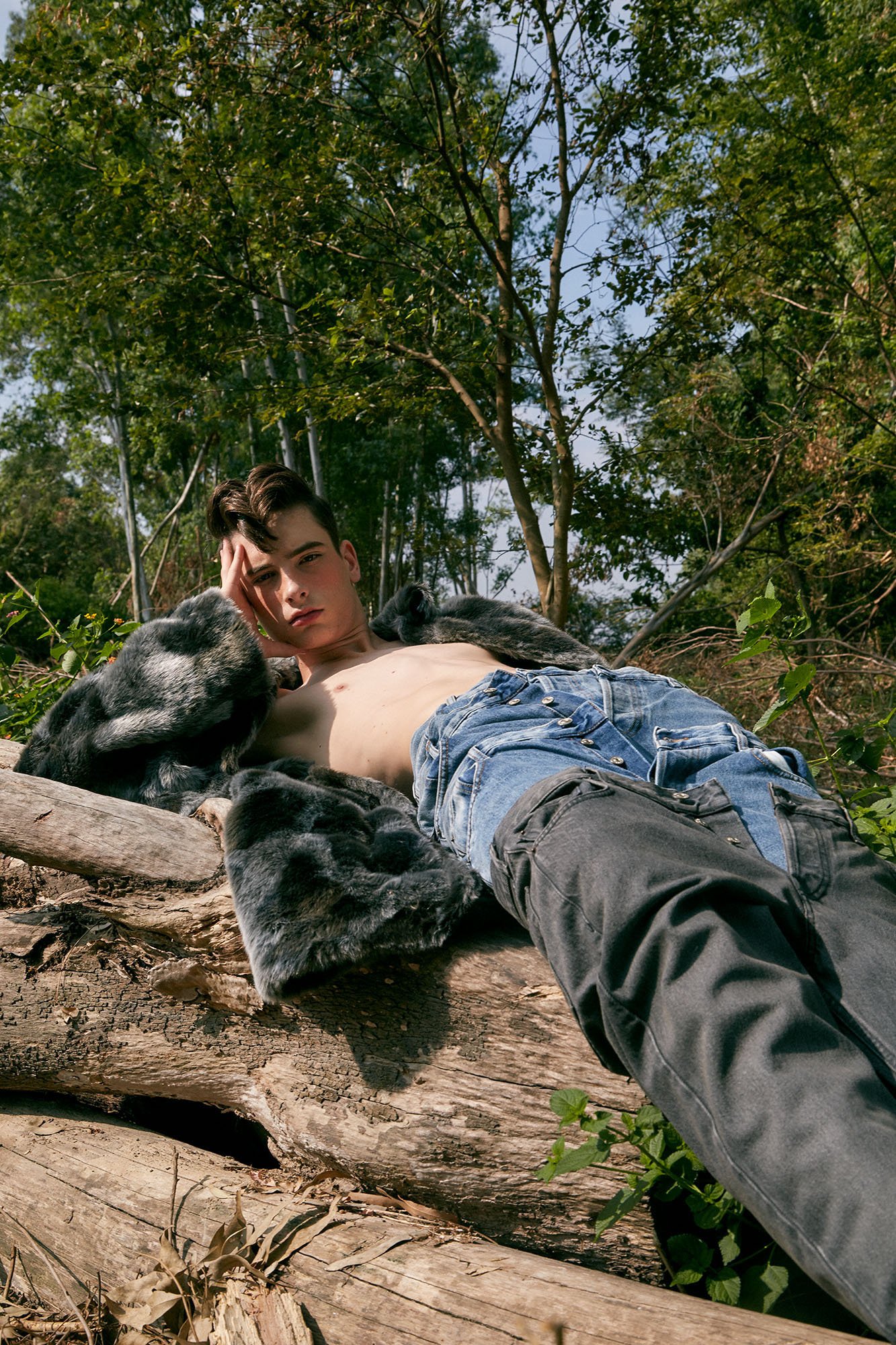 A young man lying shirtless on a fallen tree trunk in a forest, surrounded by trees and foliage, with a serious expression.