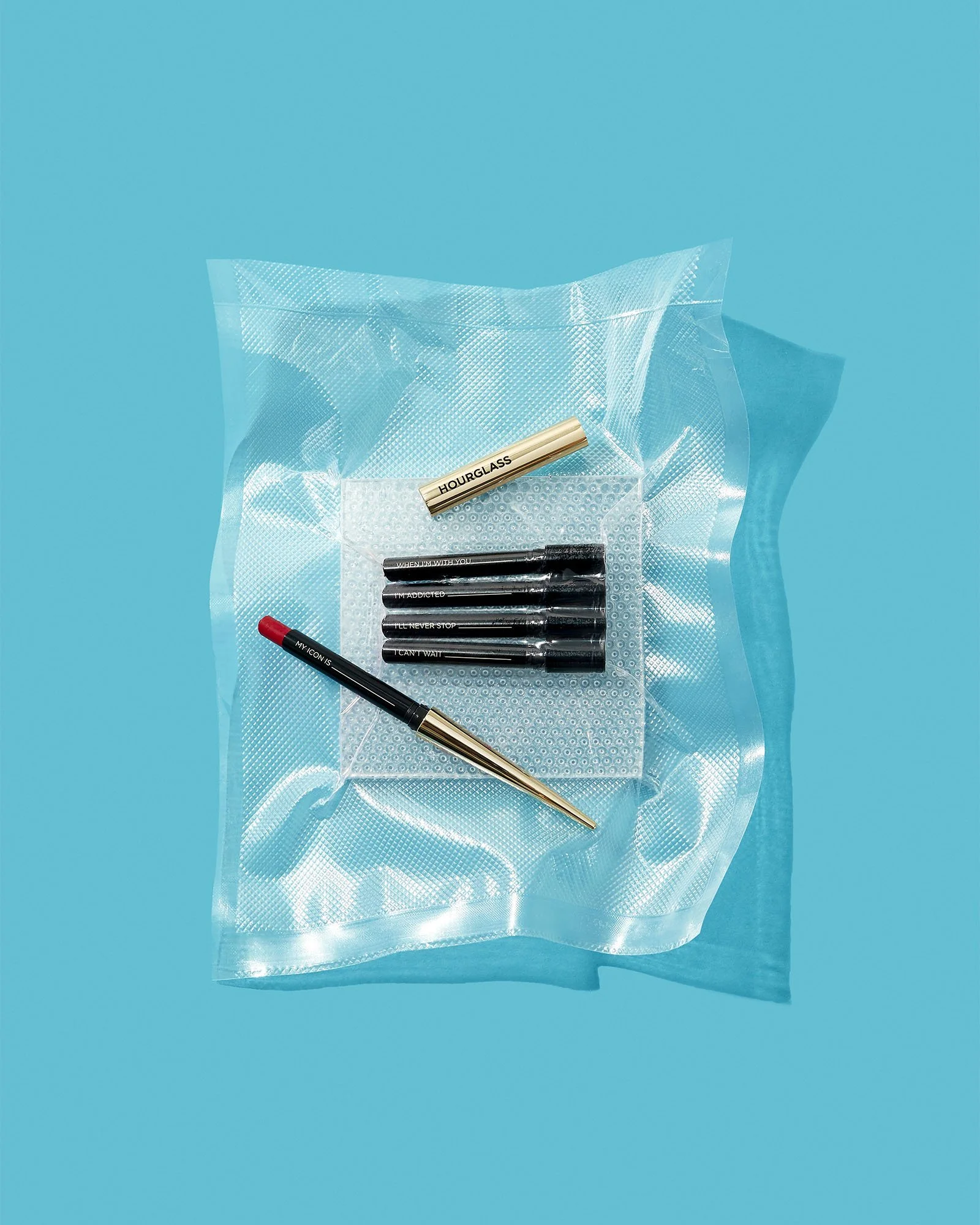 A set of black and red markers and a metallic gold pen resting on a clear, textured plastic bag on a bright turquoise background.