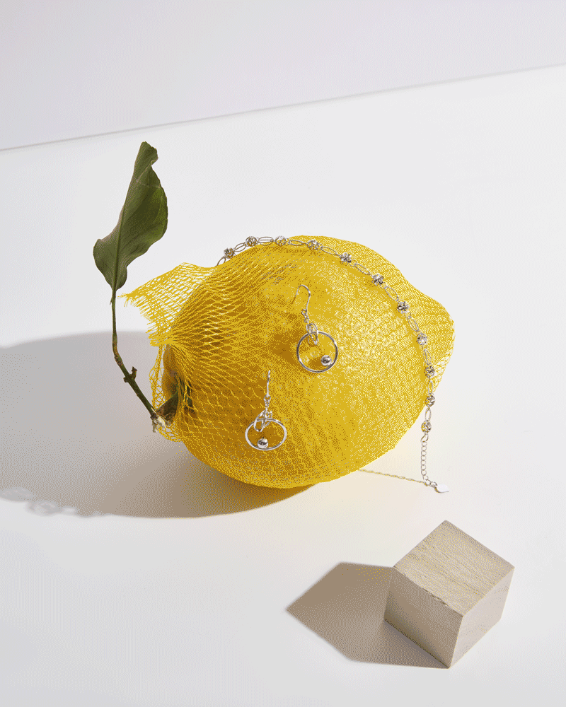 A yellow lemon wrapped in a yellow mesh bag, with a green leaf attached to its stem. The lemon has silver jewelry including earrings and a necklace on it. There is a small wooden block and a shadow cast on a white surface.