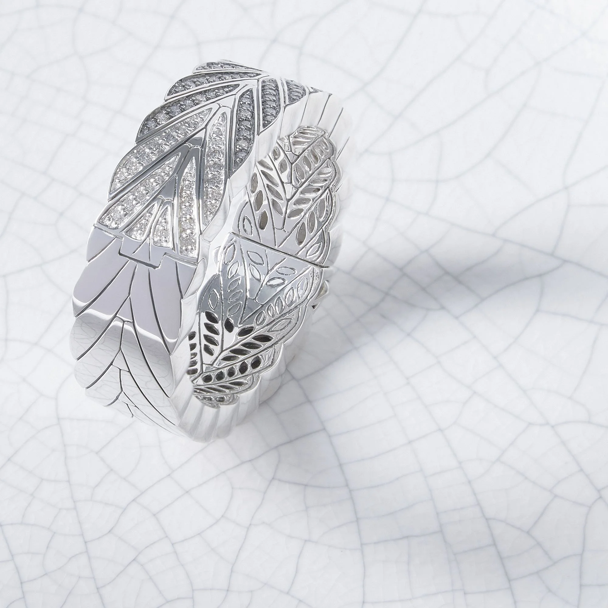 Silver ring with leaf and feather patterns, some parts decorated with small diamonds, placed on a white textured background.
