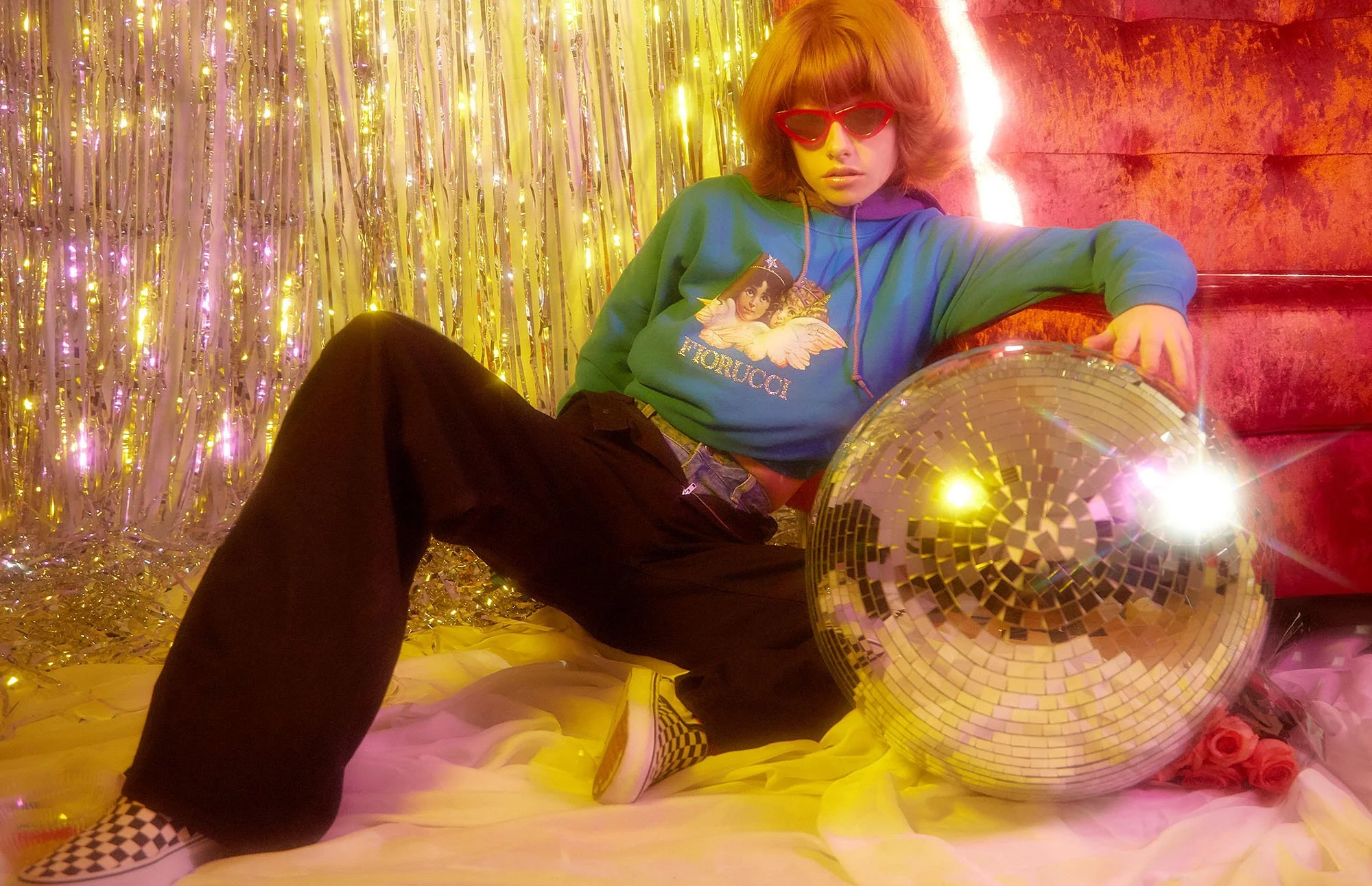 A person with short curly red hair, wearing red glasses, a blue sweatshirt with angel artwork and text 