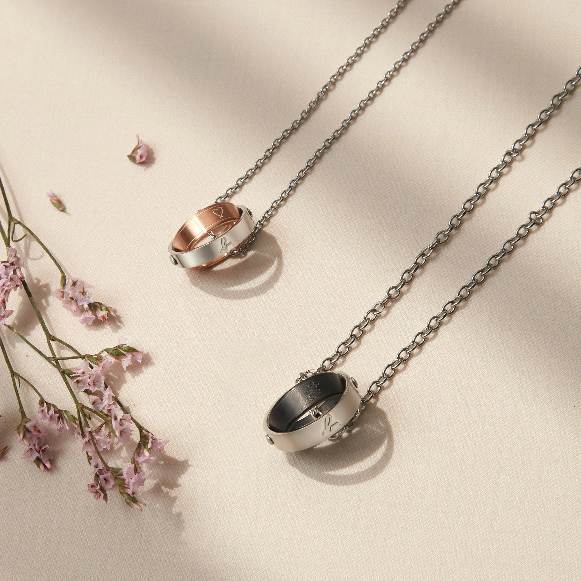 Two metallic necklaces with embedded rings on a cream fabric background, with pink flowers on the side.
