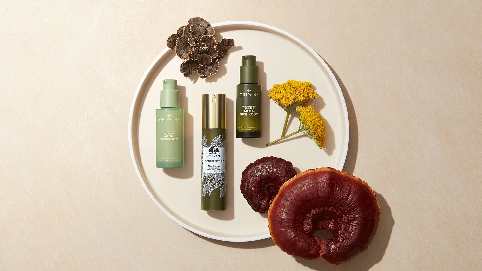 Three skincare bottles labeled "Origins Mega-Mushroom" and "Plantscription" on a white round tray, accompanied by yellow flowers, dried fungi, and a large red-brown fungus on a beige background.