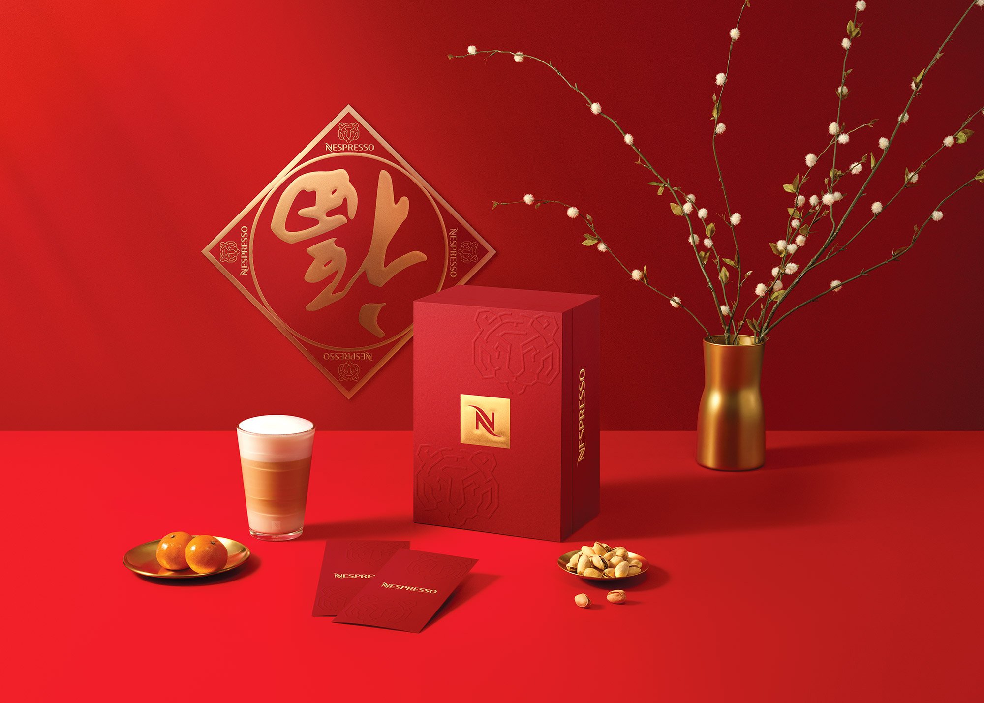 Red Nespresso coffee box, a matching card, a tall glass of layered coffee drink with foam, a small plate with two kumquats, a small bowl of pistachios, and a gold vase with white flowering branches, all set against a red background with a gold and re