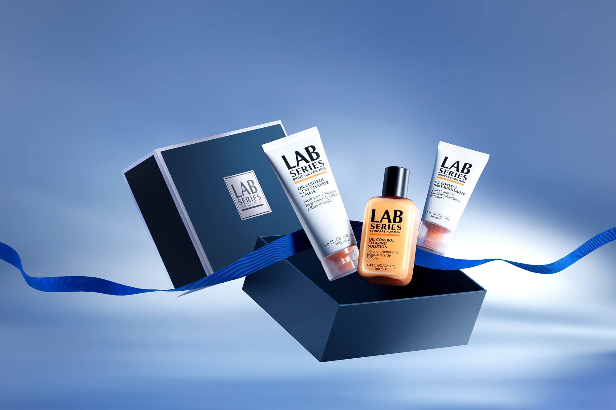 Skincare products from LAB Series for men, including a clay control cleanser, oil control moisturizer, and a solution, arranged inside a dark blue box against a light blue background with a blue ribbon.