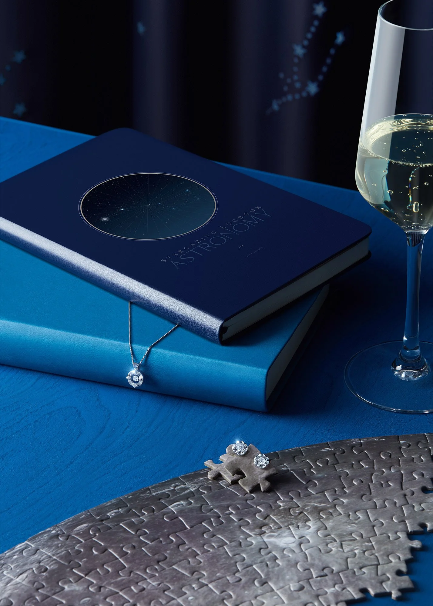 A blue astronomy-themed hardcover book titled 'Stargazing Notebook Astronomy' with a star chart on the cover, a necklace with a circular pendant resting on another book, silver puzzle pieces with rhinestone accents, a half-filled glass of champagne, 