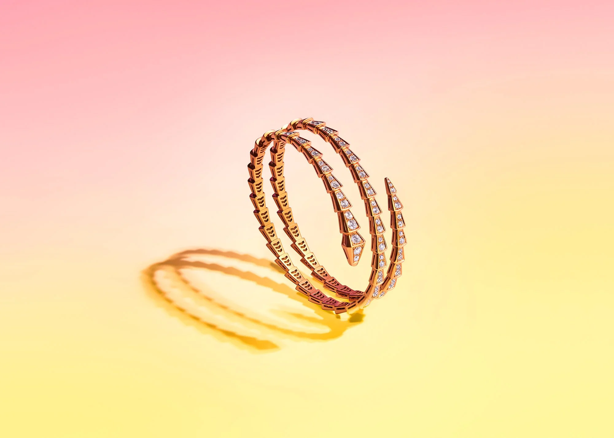Three gold bangles adorned with diamonds, casting shadows on a pastel pink and yellow background.