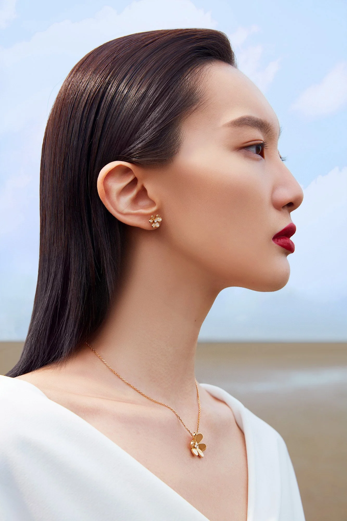 A woman with long, straight, brown hair and red lipstick wearing diamond-studded flower earrings and a butterfly necklace, standing outdoors against a cloudy sky.