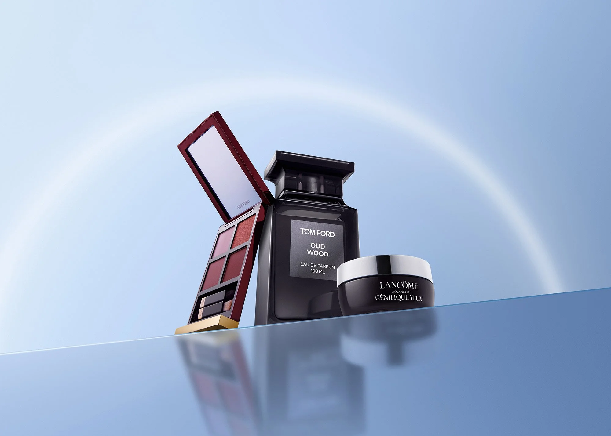 Arrangement of luxury cosmetic products including a lipstick, perfume, and eye cream on a reflective surface with a blue background.