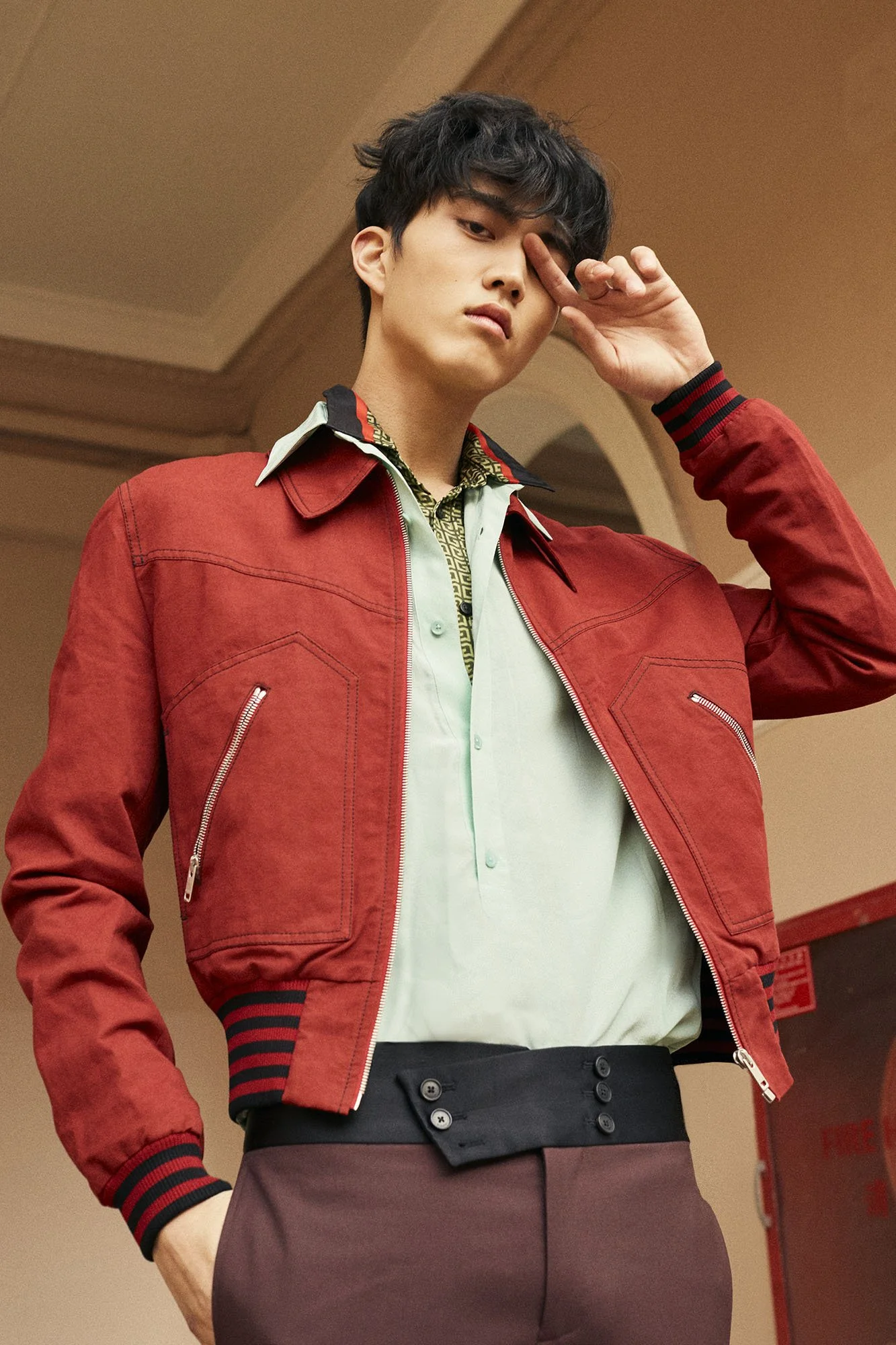 A young man wearing a red jacket, light green shirt, and maroon pants, posing indoors with one hand touching his face.