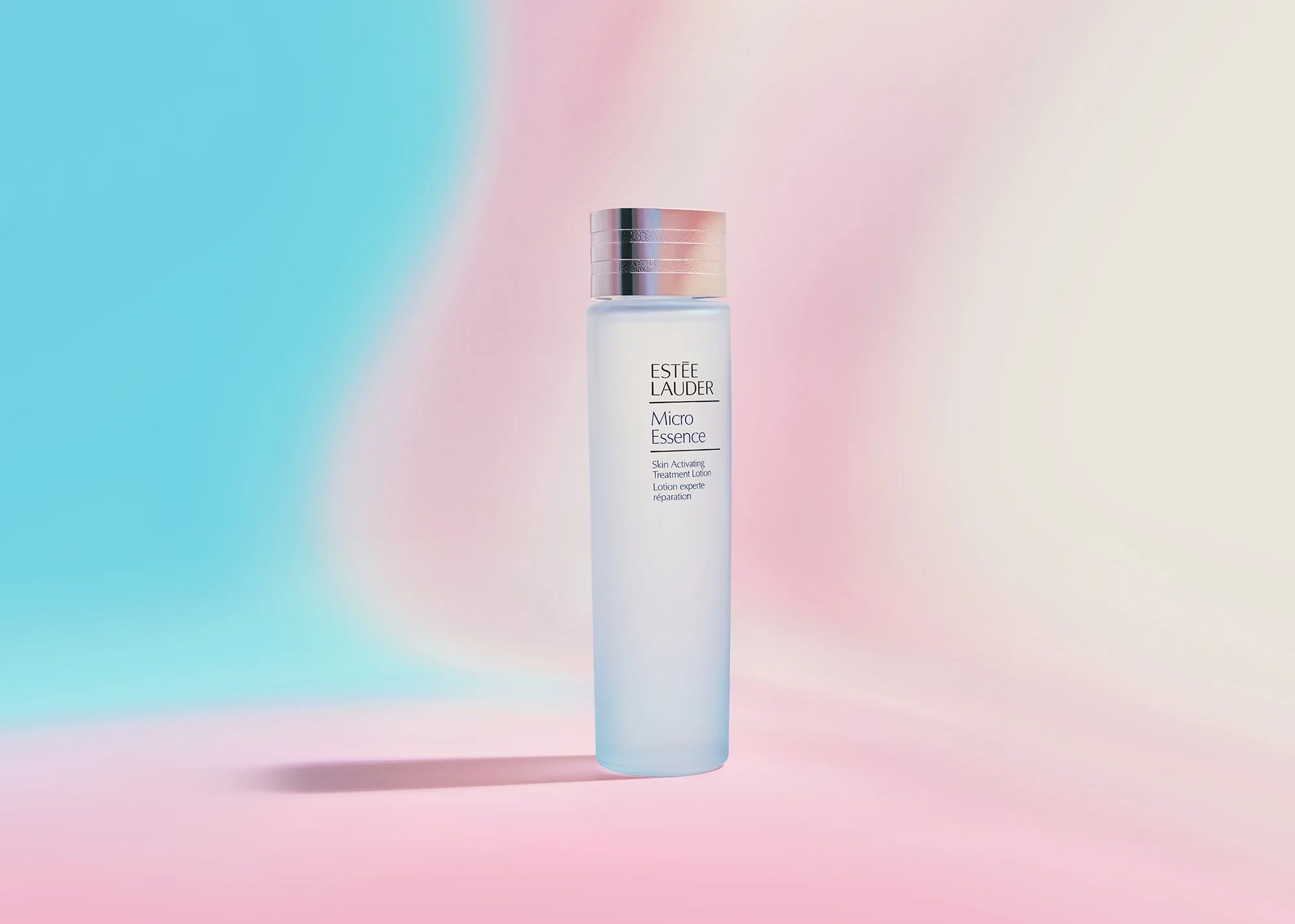 A bottle of Estée Lauder Micro Essence Skin Activating Treatment Lotion standing on a pastel-colored background with pink, blue, and white gradients.