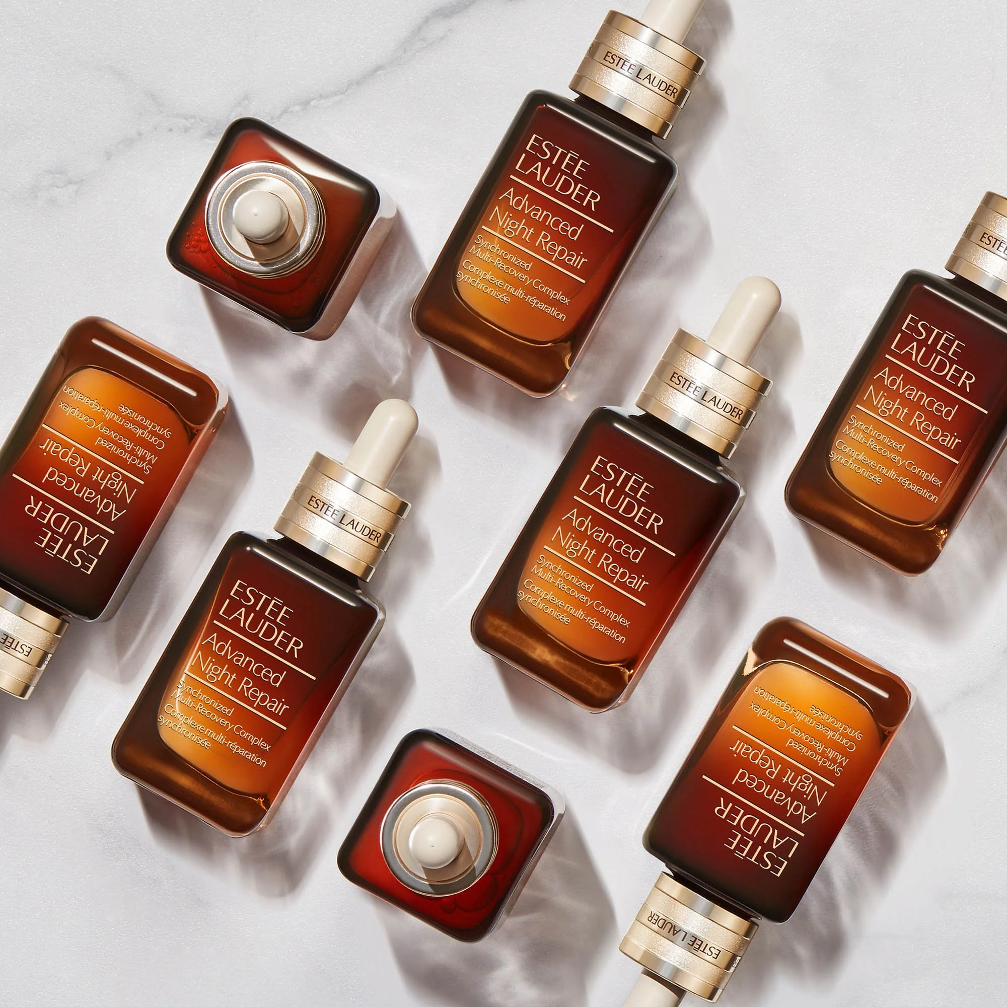 Multiple bottles of Estée Lauder Advanced Night Repair serum arranged on a white marble surface, some bottles are upright and others are lying down.