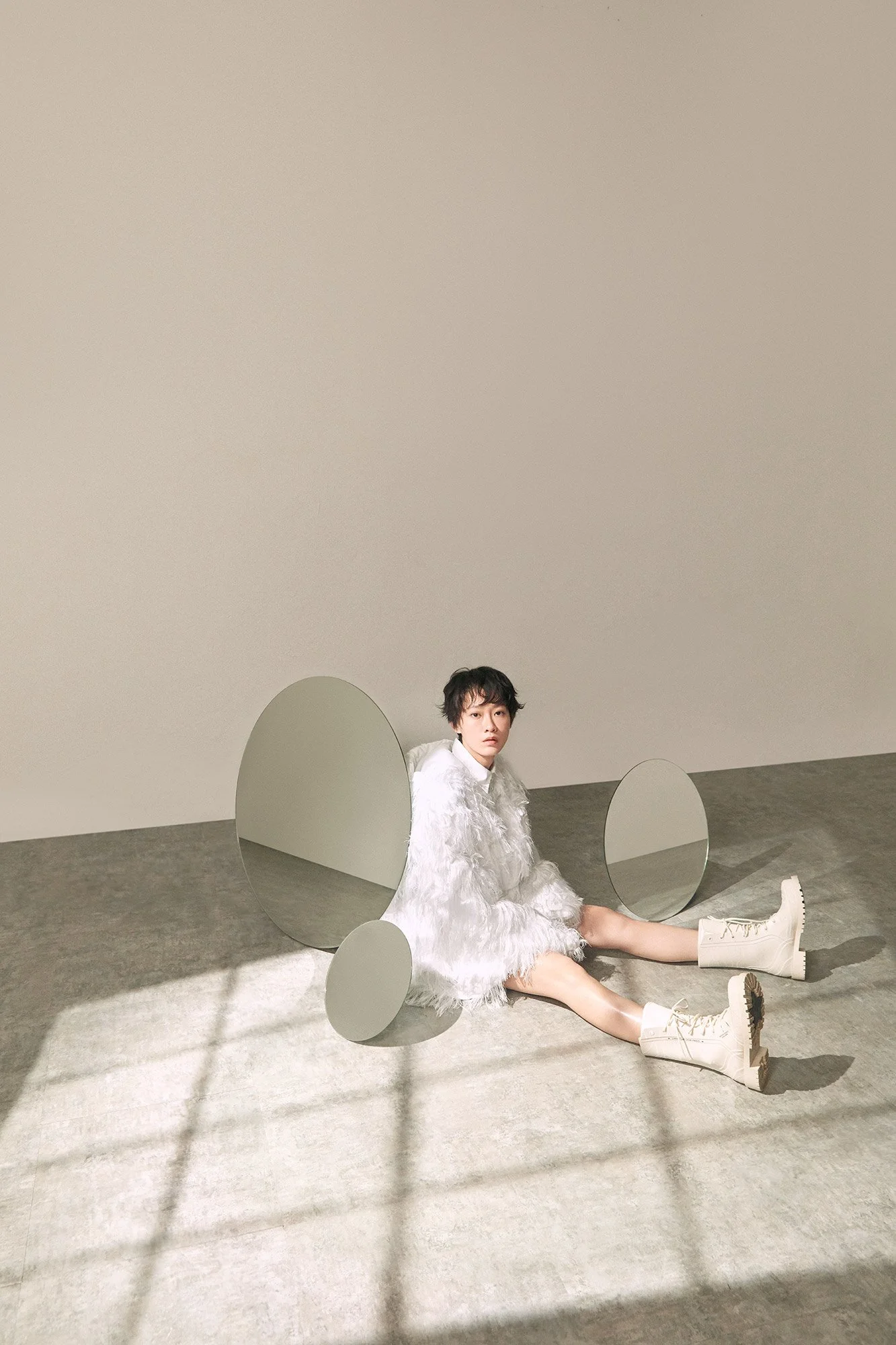 A woman in white fuzzy clothing with beige boots sits on the floor against a beige wall. There are three round mirrors on the floor reflecting parts of her. Shadows and sunlight create a grid pattern on the carpet.