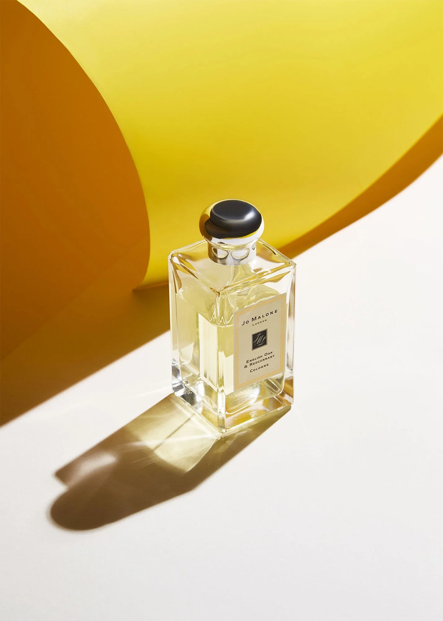 A clear glass perfume bottle with a black cap on a white surface, casting a shadow, with a large yellow and orange abstract shape in the background.