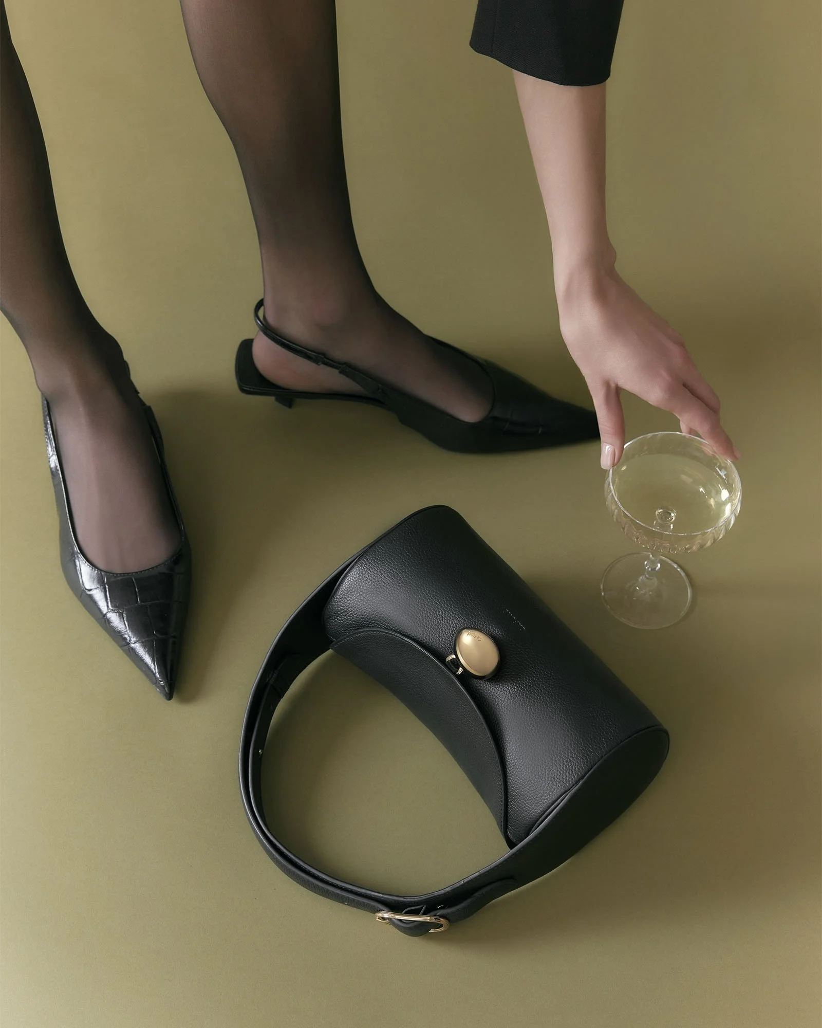 A black purse with a gold clasp, a glass cup with a drink, a pair of black shoes with a small heel, and a person wearing sheer black stockings and black shorts, reaching for the glass.