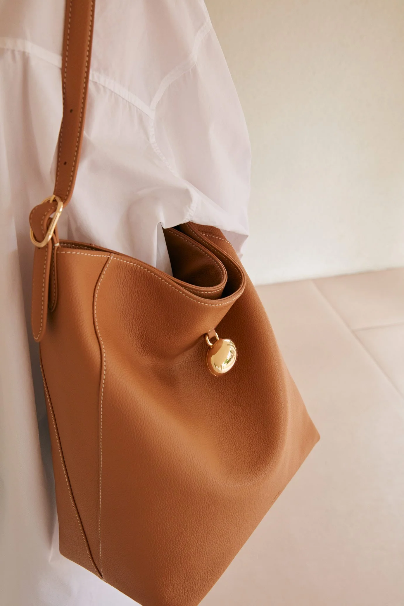 A tan leather handbag with gold hardware hanging from a white shirt on a white wall.