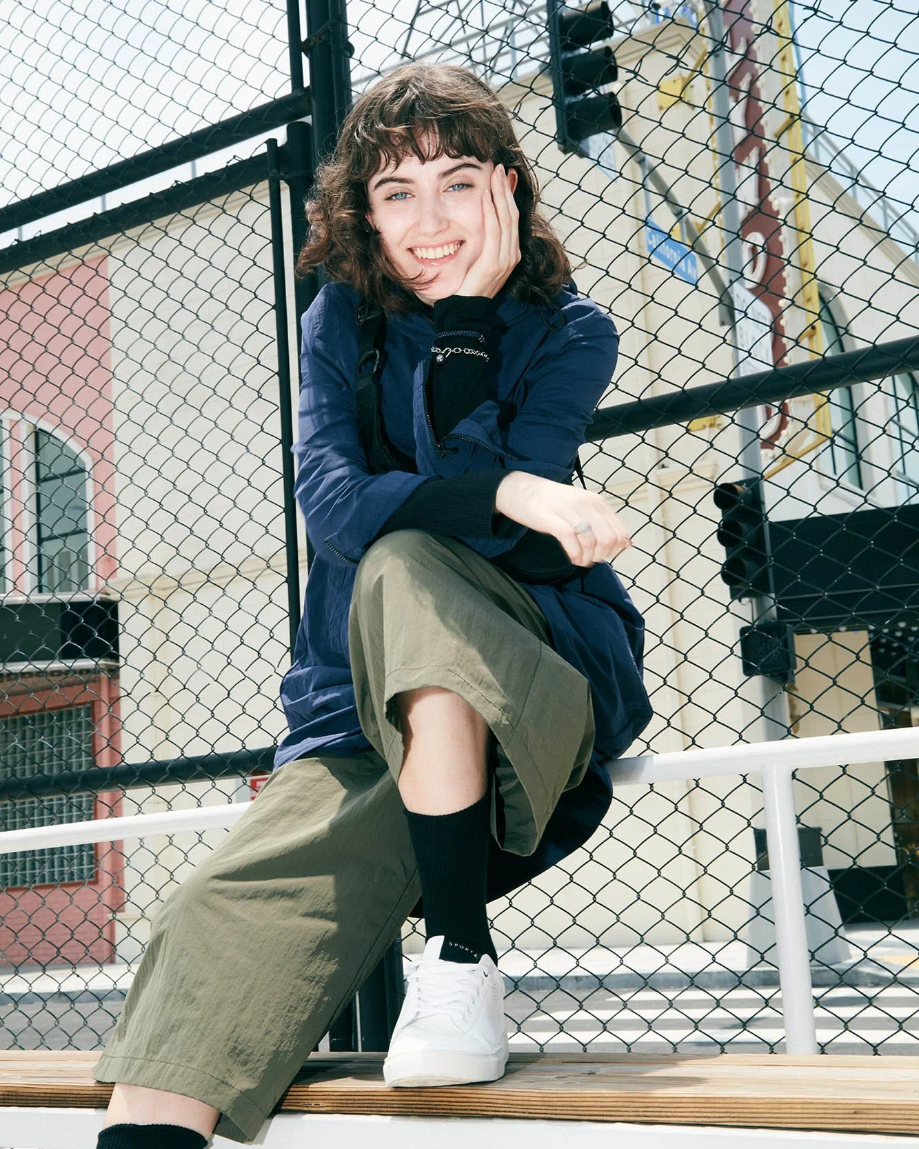 A young woman sitting on a bench behind a chain-link fence, smiling, with a city building and fire escape in the background.