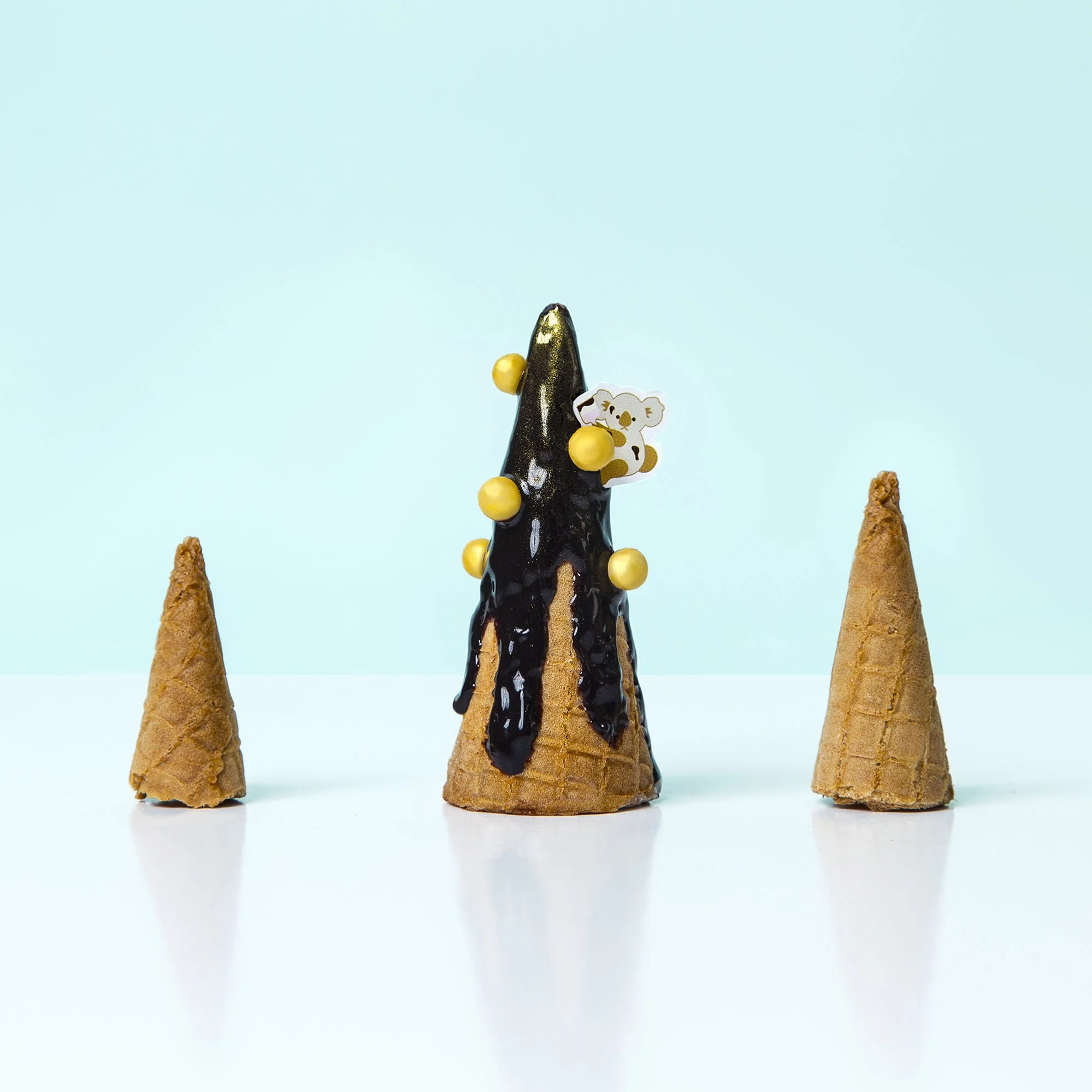 Three ice cream cones arranged like a birthday cake with a koala decoration, topped with black chocolate glaze and gold decorative balls, against a light blue background.