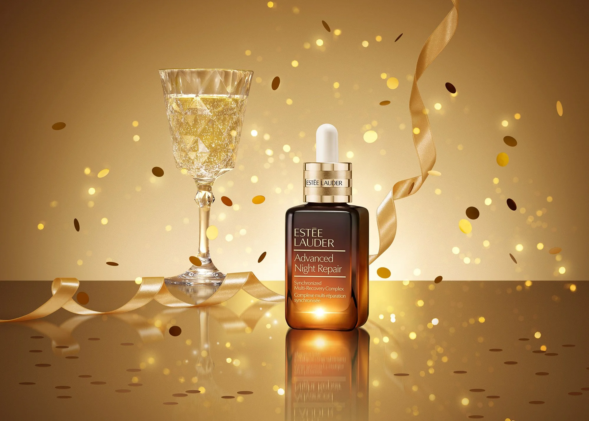 A bottle of Estée Lauder Advanced Night Repair serum on a reflective gold surface, surrounded by gold confetti and a gold ribbon, with a wine glass filled with champagne in the background.