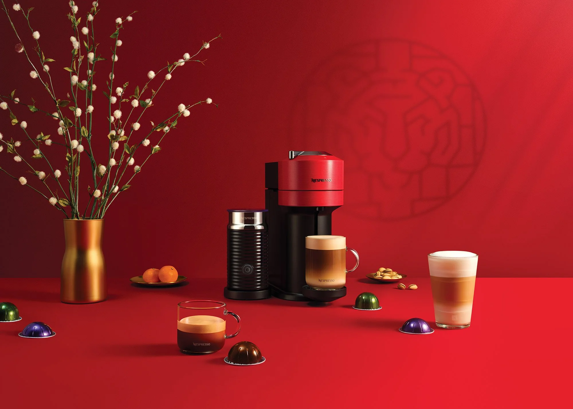 Red Nespresso coffee machine with cups of coffee, chocolates, and a vase with flowers on a red table and wall.