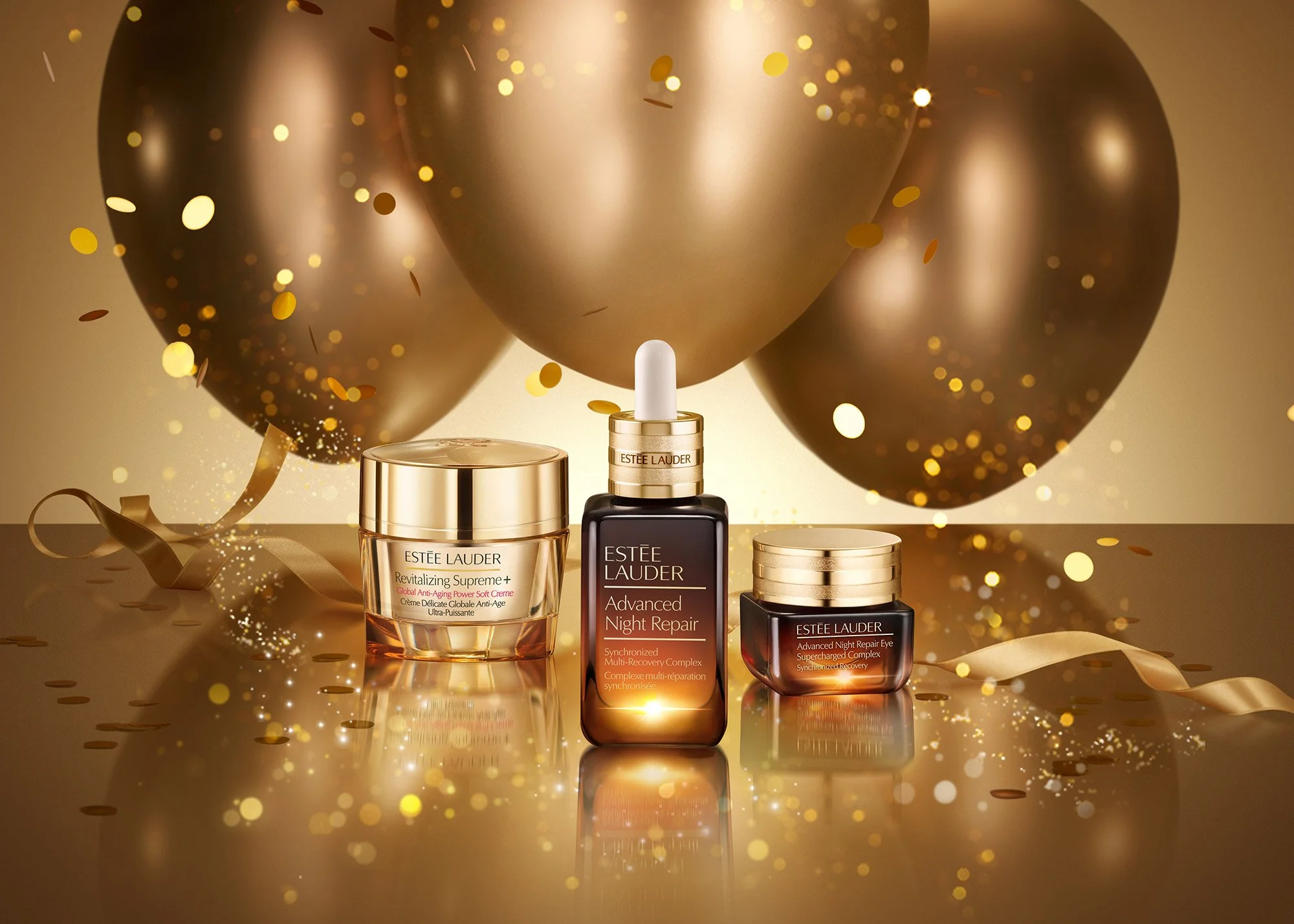 Estée Lauder skincare products displayed with black and gold balloons and gold confetti, including Revitalizing Supreme+ cream, Advanced Night Repair serum, and Advanced Night Repair Eye cream, on a reflective surface.