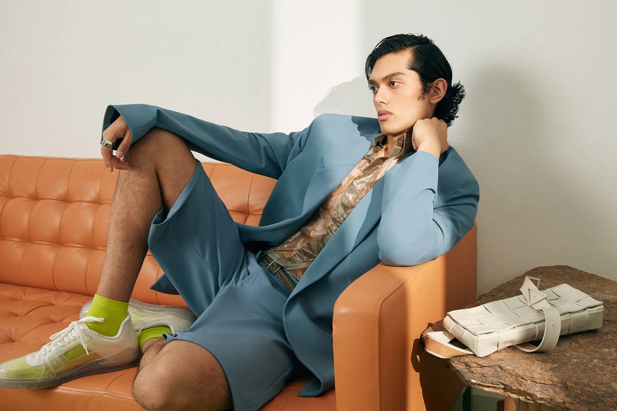 A young man with dark hair and light skin lounging on an orange leather sofa. He is wearing a blue suit with a patterned shirt, bright green socks, and white sneakers. His left leg is resting on the sofa, and he has a ring on his right hand. There is