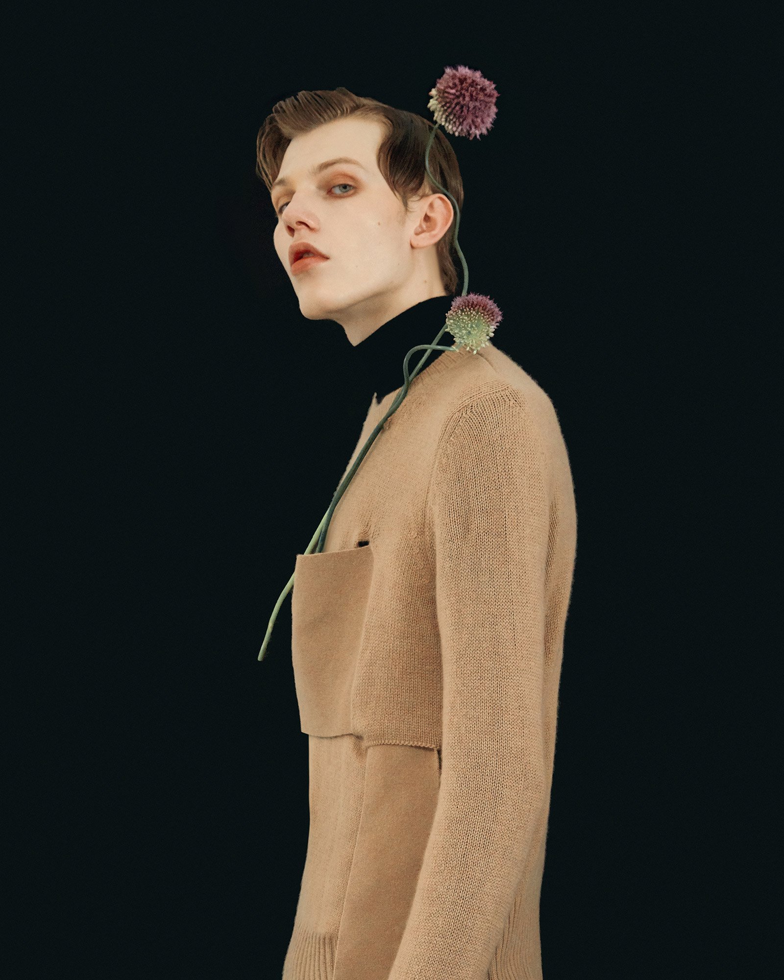 A young man with light skin and short, styled hair wearing a black turtleneck and a beige coat, with purple flower stems around his neck and on his head, against a dark background.