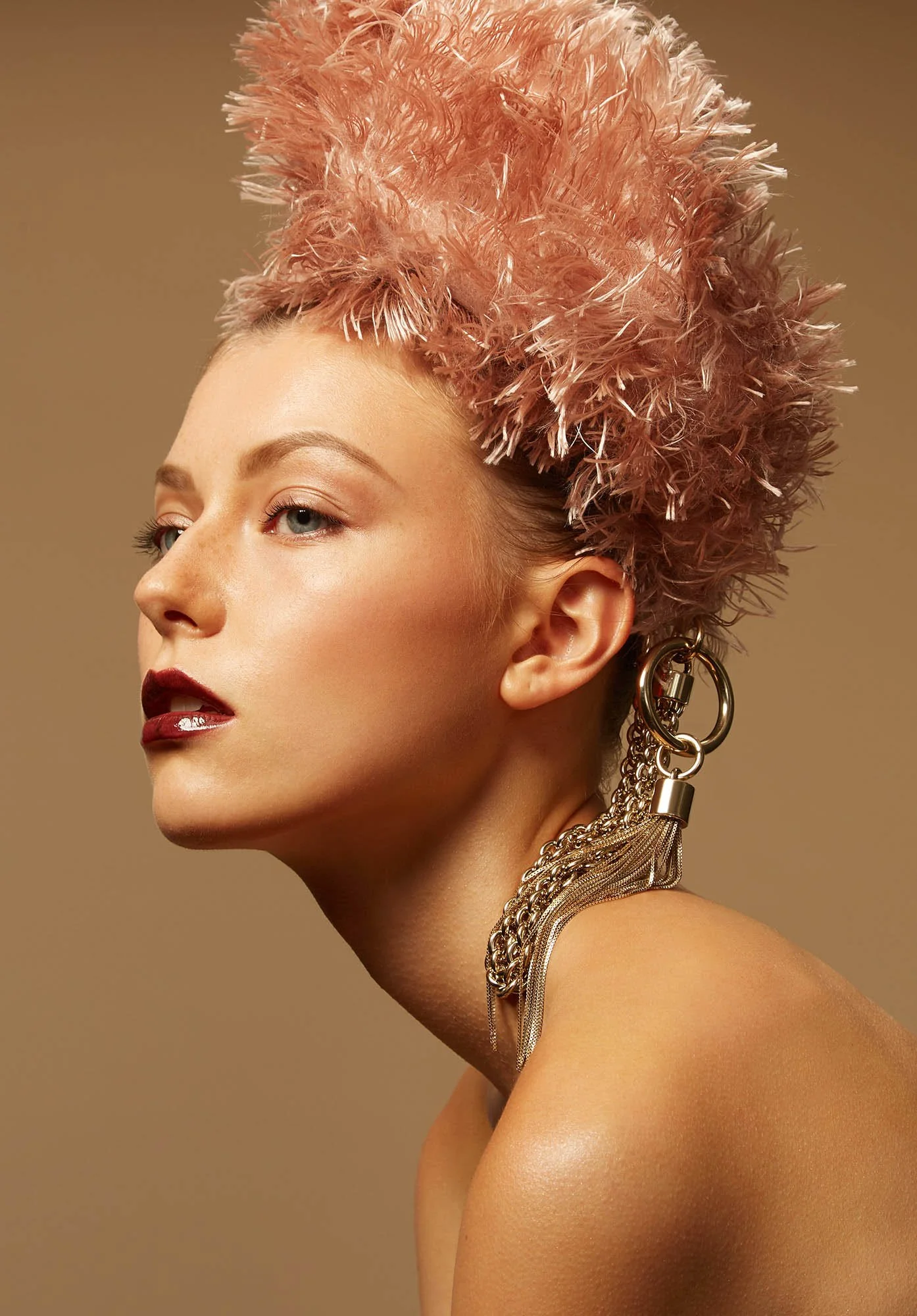 A woman with pink feathered hair, wearing large statement earrings and a chain necklace, against a neutral background.