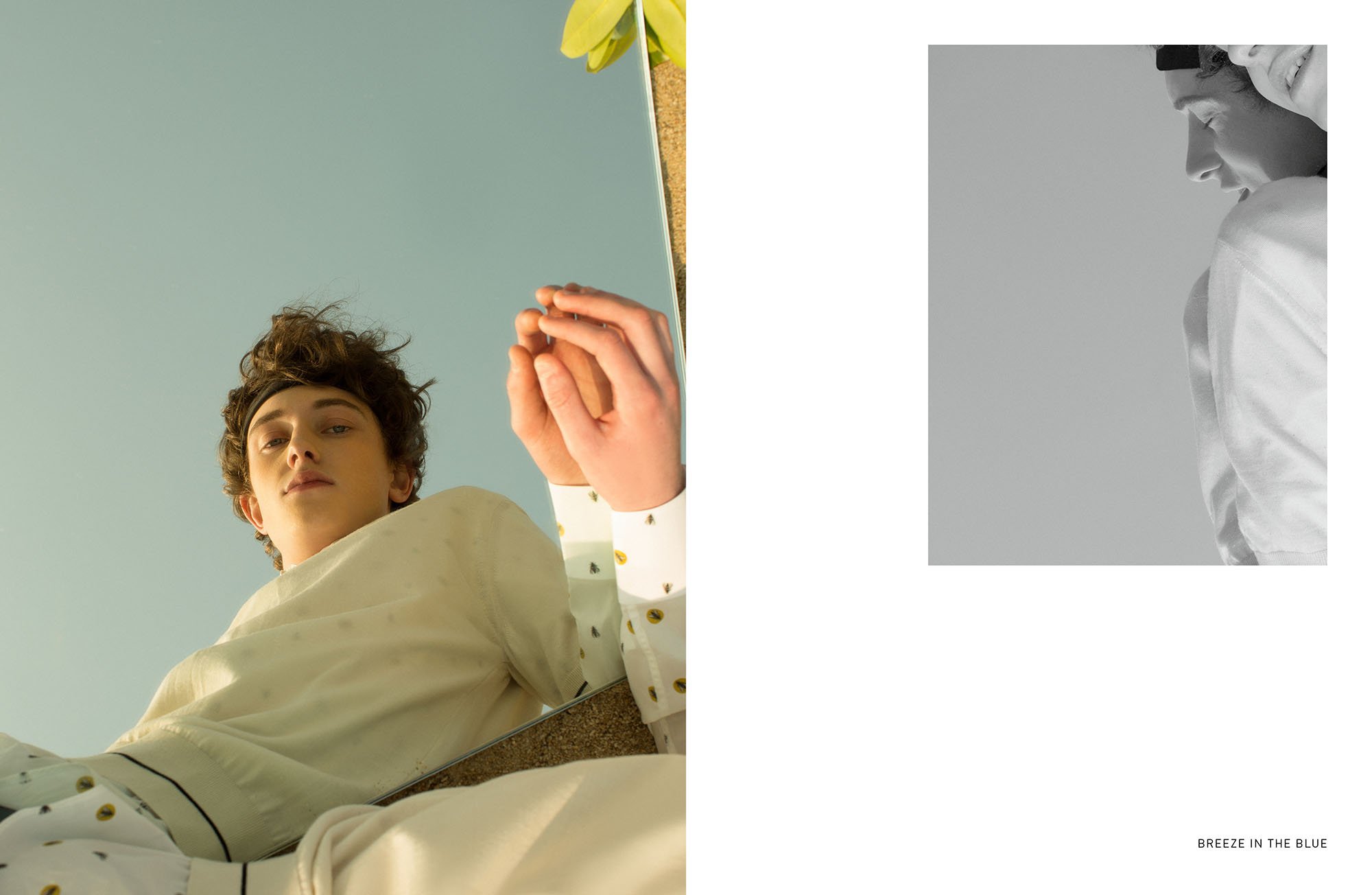 A young person with curly hair lies on the ground, looking up at the camera through a mirror, with a light blue sky above. The person wears a light-colored, long-sleeve shirt with small yellow and black patterns. To the right, there is a small black-